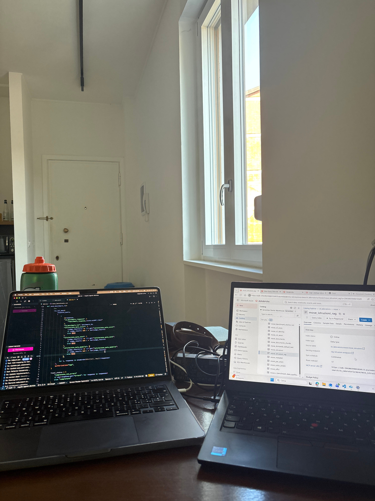 The image shows a desk with two laptops, one open showing code and the other displaying a web interface. A green water bottle with an orange cap is positioned to the left of the laptops. In the background, a white door with gold hardware is visible, and a window with natural light streaming through it is to the right. The overall setting appears to be an indoor workspace, possibly a home office or a co-working space. There are no people visible in the image. The environment suggests a focused work session.