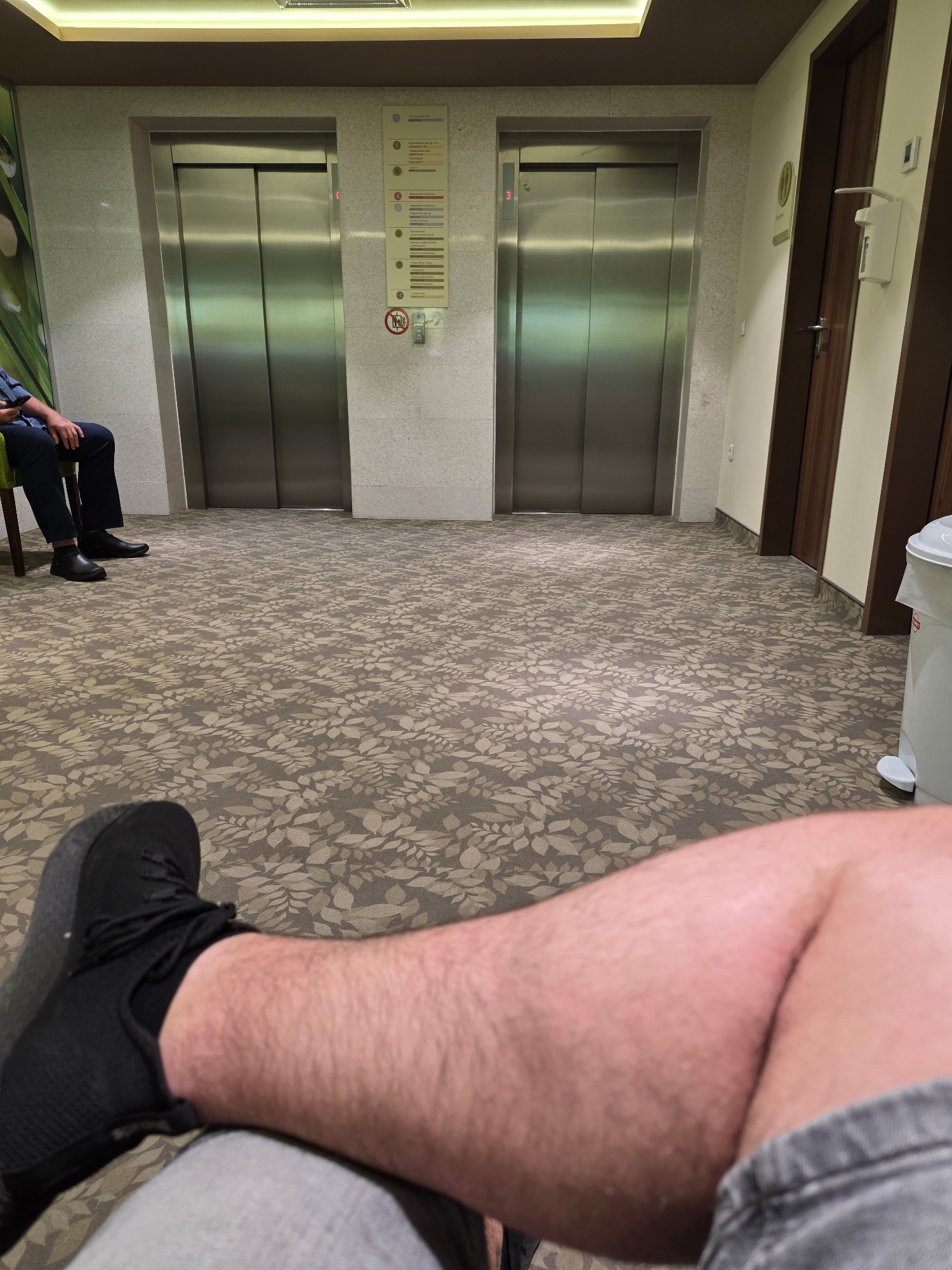 The image depicts an indoor scene with two elevators side-by-side, both with their metallic doors closed. To the left, a person is seated on a chair, wearing dark trousers and black shoes, and holding a smartphone. The floor is covered with a patterned carpet featuring a repeating leaf motif. To the right, there's a wooden door with a dark frame and a white hand sanitizer dispenser mounted on the wall above it. A white trash bin with a pedal is also visible on the far right. The walls are light-colored with some textured panels. The scene appears to be a waiting area, possibly in a hospital or an office building, given the signage and the overall clean and functional aesthetic. The location of the city of Sofia, Bulgaria is not directly discernible from the visual elements within the image itself. The foreground is dominated by the leg and foot of the person taking the photograph, wearing a black athletic shoe and grey shorts.