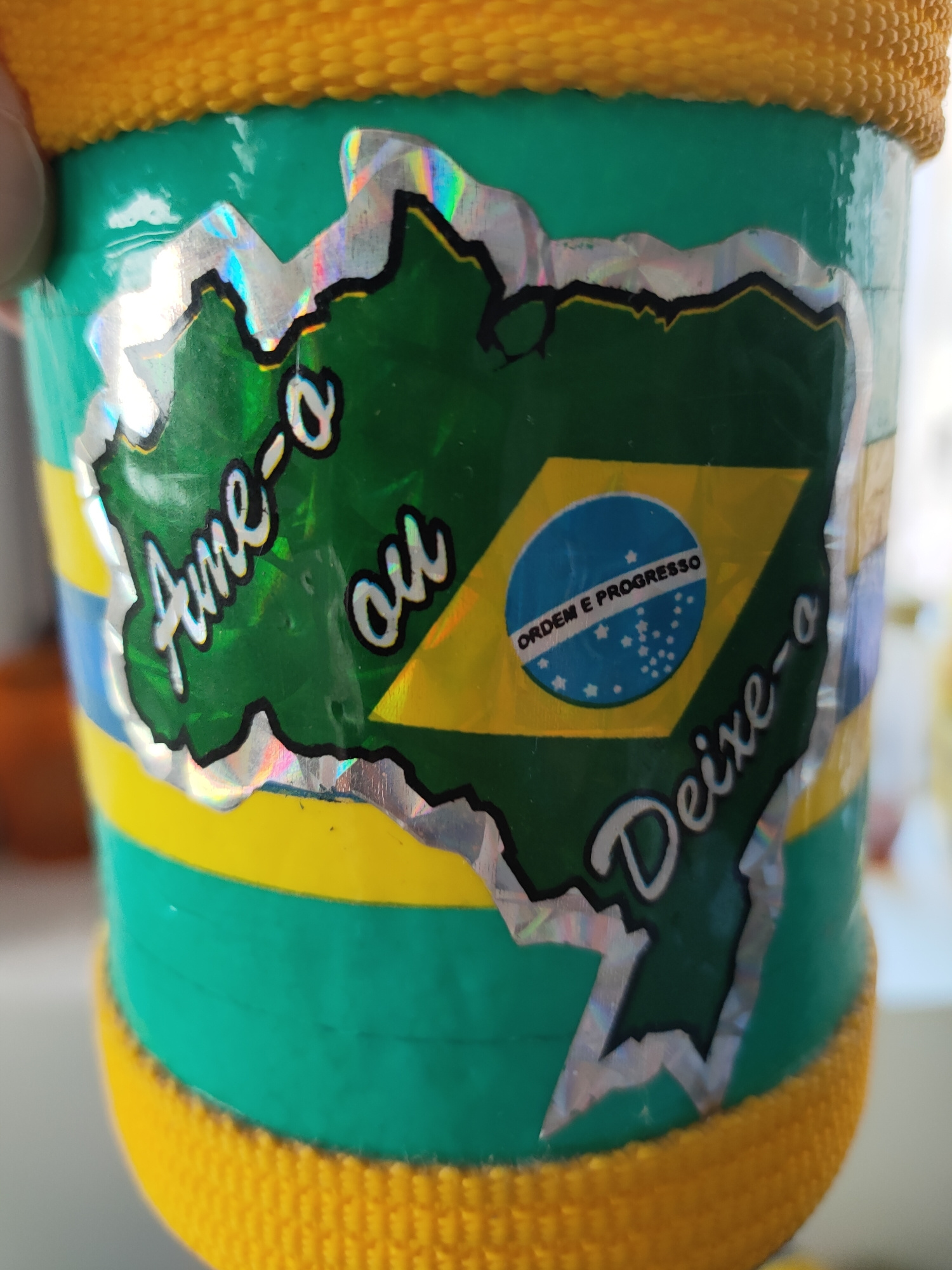 A close-up image shows a green cylindrical object, possibly a can or a bottle, adorned with a sticker featuring a stylized map of Brazil. The sticker also includes the Brazilian flag, with the motto "ORDEM E PROGRESSO" (Order and Progress) visible on the blue sphere. Text on the sticker reads "Ame-o ou Deixe-o" (Love it or Leave it). The object has bands of yellow and blue, and is topped and bottomed with a textured yellow material. The background is blurred and appears to be an indoor setting. No specific location in Perpignan, France, can be identified from the image.