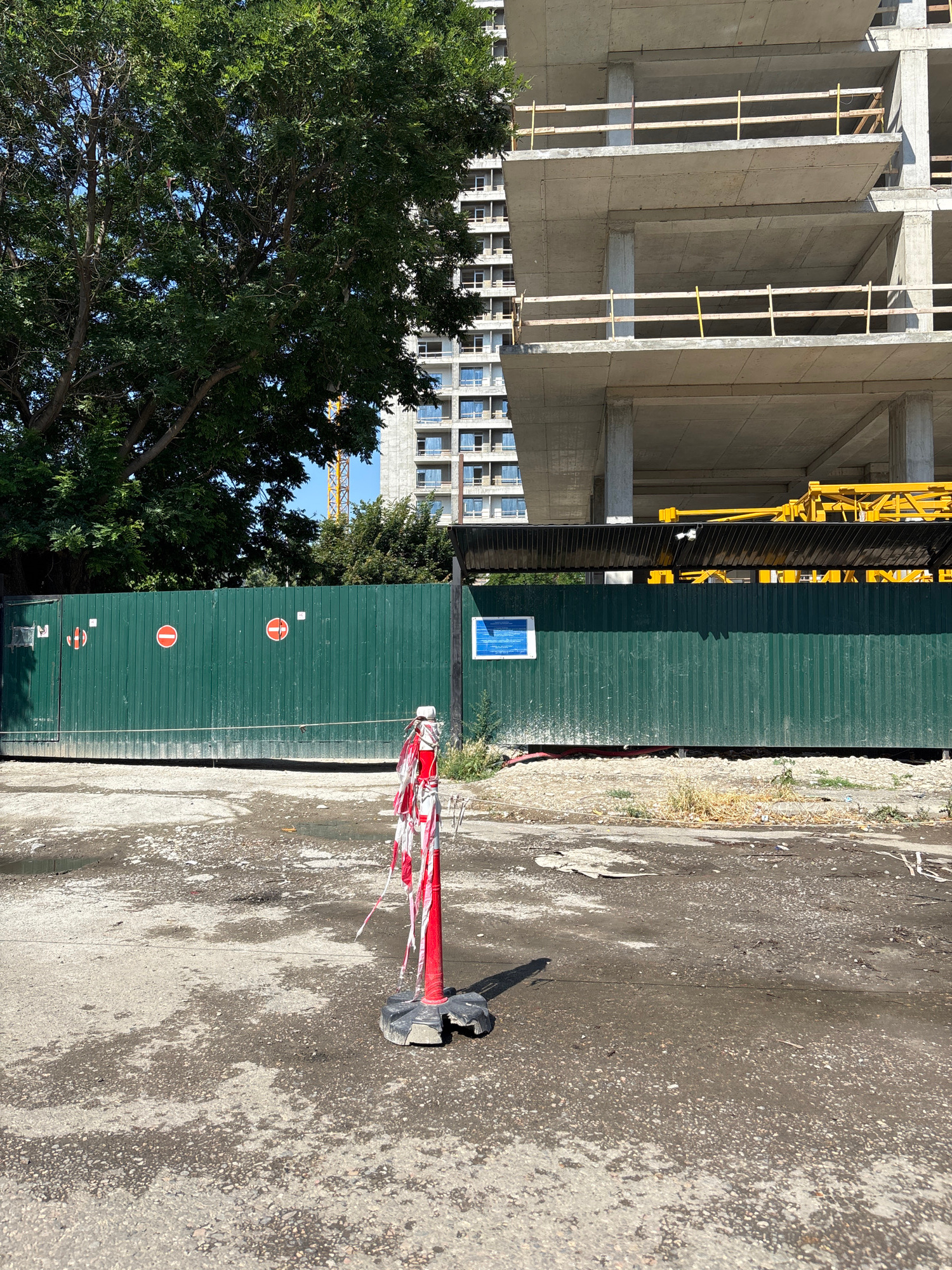 The image depicts a construction site with a partially built concrete structure in the background. A large green metal fence runs across the midground, with several "no entry" signs visible on it. To the left, a lush green tree stands tall, partially obscuring another building with multiple windows. In the foreground, a red and white striped traffic cone with a black base sits on a rough, wet-looking asphalt surface. A yellow construction crane is partially visible above the fence. The scene suggests an active construction area, likely in an urban environment, possibly Tbilisi, Georgia, given the context of the prompt, although no explicit identifying markers confirm this location. The lighting indicates it is daytime, with sunlight casting shadows.