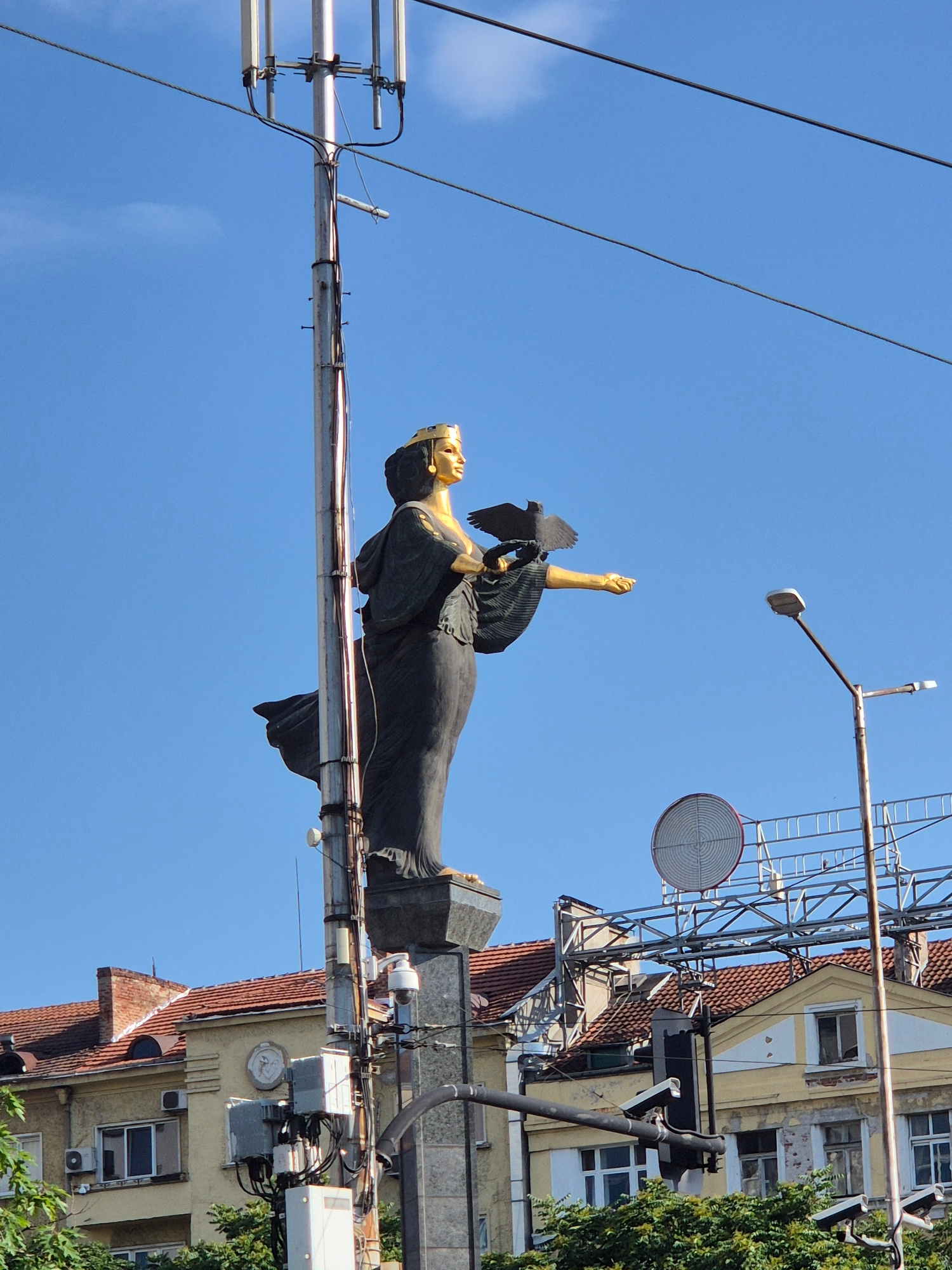 This is a medium-angle, eye-level shot of the Saint Sofia statue in Sofia, Bulgaria. The statue, made of bronze and gilded elements, depicts a female figure wearing a golden crown, a dark robe, and holding a raven in her left hand while her right arm is extended. The statue stands on a stone pedestal atop a metallic pole that supports telecommunication antennas and various cables. In the background, a clear blue sky is visible with a few clouds. Below the statue, the rooftops of buildings, some with red tiles, are seen. A streetlight and a circular radar-like structure are also present in the mid-ground, along with more antennas and wiring. The overall environment suggests an urban setting with modern infrastructure amidst historical architecture.