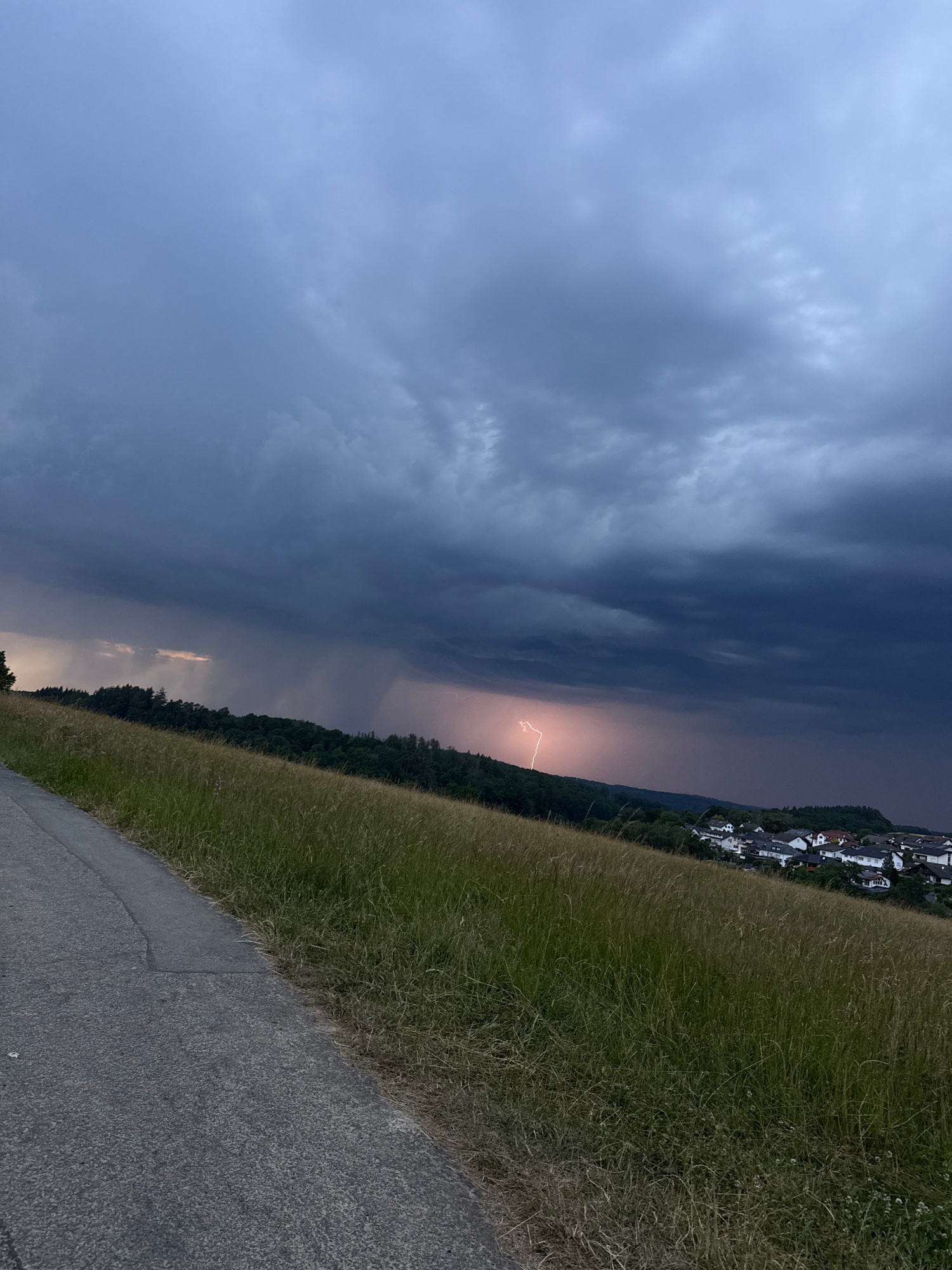 An outdoor, medium shot captures a dramatic twilight scene with a thunderstorm overhead. The foreground features a paved path curving to the left, bordering a tall, dry grass field. Beyond the field, a line of dark green trees stretches across the middle ground, leading to a small village nestled on a hillside in the background. The sky is filled with dark, ominous storm clouds, punctuated by a vibrant flash of lightning striking somewhere over the village. Rain can be seen falling in the distance. The overall atmosphere is one of impending or ongoing inclement weather. The location is identified as Oberquembach, Germany.