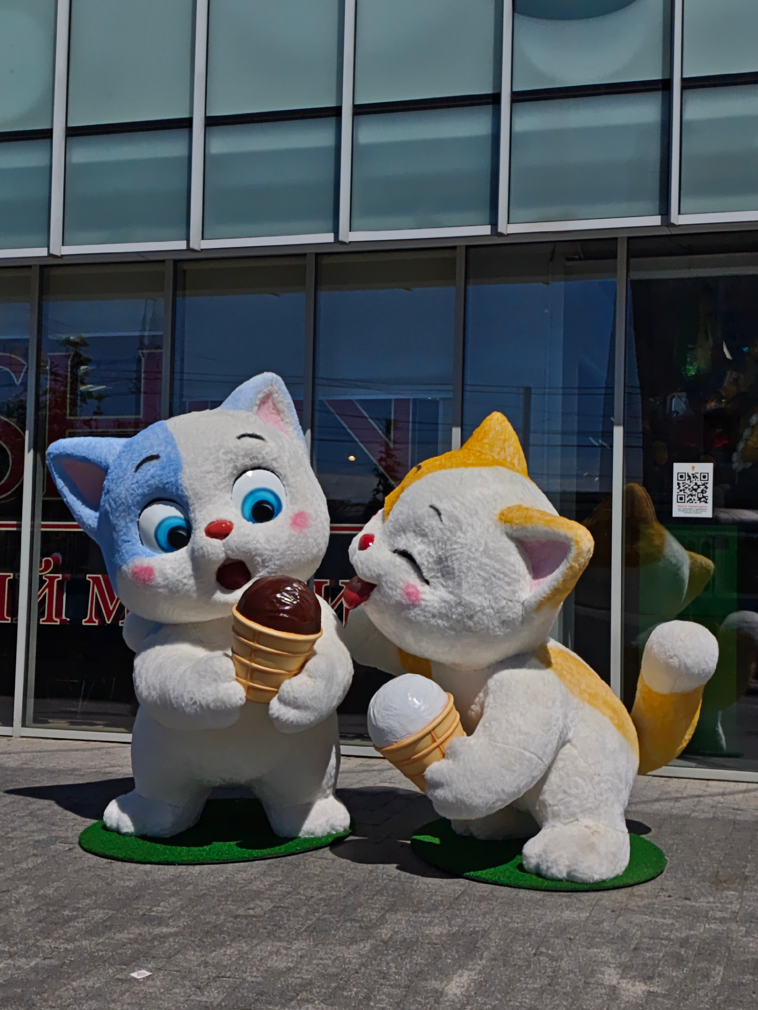 This image depicts two large, plush cat statues placed outdoors on a paved surface in front of a building with a glass facade. The statue on the left is predominantly white with blue markings on its head and ears. It has large, round blue eyes, a small red nose, and pink cheeks. This statue is holding a chocolate ice cream cone in its paws, with the cone angled towards its mouth. The statue on the right is white with orange markings on its head and ears. It has closed eyes, pink blush marks on its cheeks, and its mouth is open as if licking. This statue is holding a vanilla ice cream cone. Both statues are standing on small green circular mats that resemble grass. In the background, the building's glass windows reflect the sky and surroundings. A QR code is visible on one of the windows. The overall scene suggests an outdoor decorative installation, possibly outside a business or entertainment venue. The location is identified as Vinnytsia, Ukraine.