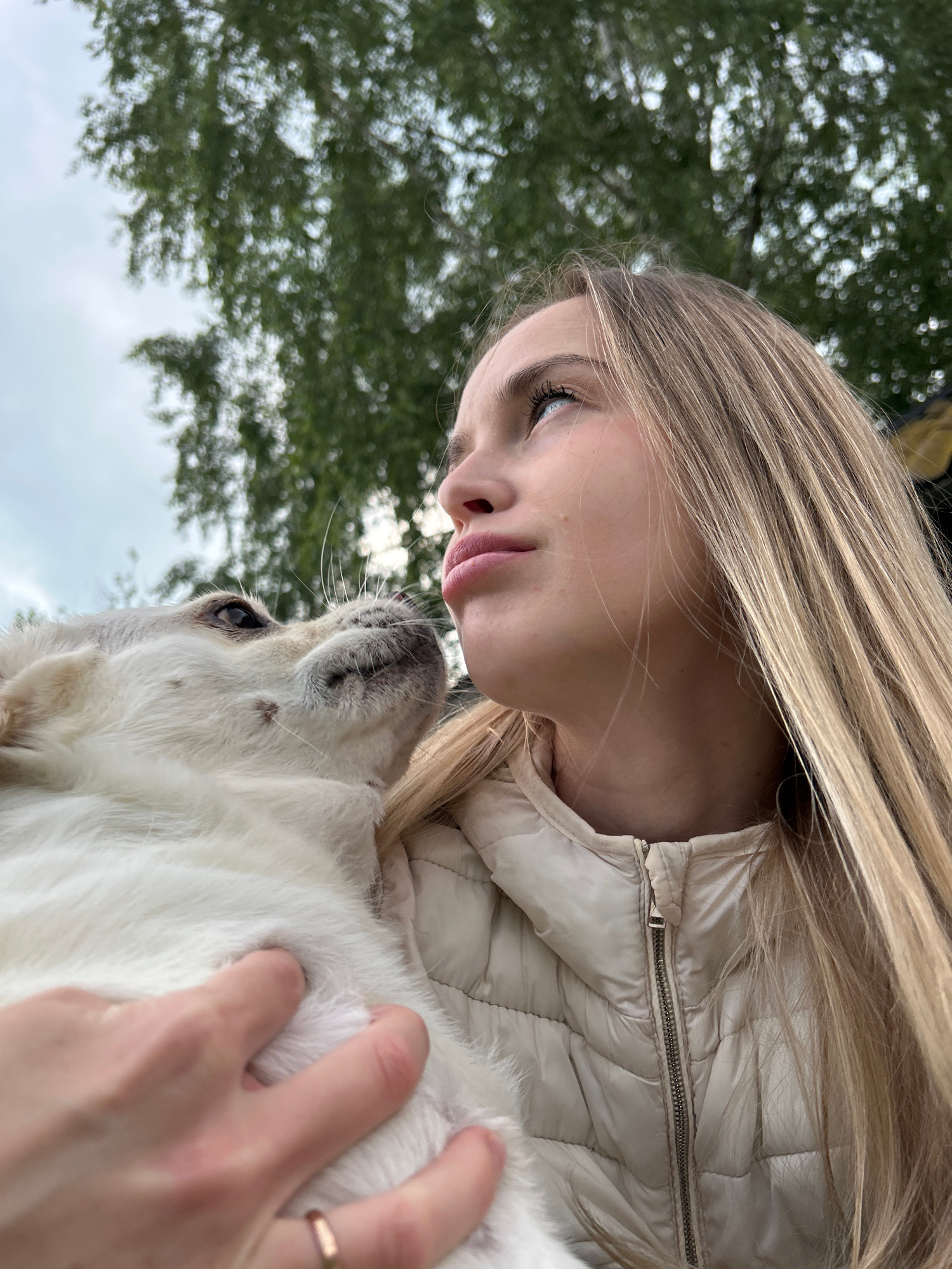 A young blonde woman with long, straight hair is holding a white puppy to her face, looking upwards. The woman is wearing a light-colored puffer jacket. The puppy's snout is close to her lips, and the dog's eyes are directed towards the woman. The background shows out-of-focus green foliage and a cloudy sky, suggesting an outdoor setting. The image was taken in the city of Азинский, Russia.