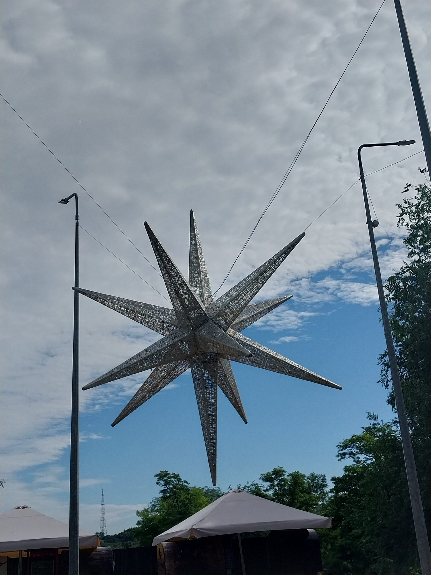 A large, metallic, star-shaped sculpture with multiple points hangs suspended from wires against a sky with scattered clouds. Two streetlights, one on the left and one on the right, are visible in the frame. In the background, a forest of green trees can be seen, and further in the distance, a transmission tower is visible on a hill. At the bottom of the image, parts of two white umbrellas and some wooden structures are present, suggesting an outdoor seating area or market. The overall scene is an outdoor public space with decorative elements.