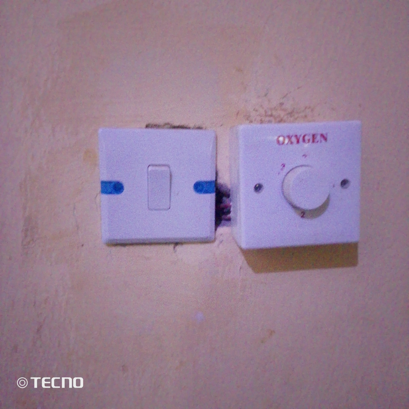 Two electrical wall plates are mounted on a textured, light-colored wall. The plate on the left is a white light switch with a blue toggle. The plate on the right is a white dial control with the word "OXYGEN" printed in red text above a circular dial with numerical markings. The dial appears to be set to a low number, possibly "1" or "2". The overall lighting is dim, and the image has a grainy quality. A watermark in the bottom left corner reads "TECNO". The scene is likely an indoor electrical fixture in a residential or commercial setting. Based on the context provided by the query about Jalingo, Nigeria, this image was taken in Jalingo, Nigeria.