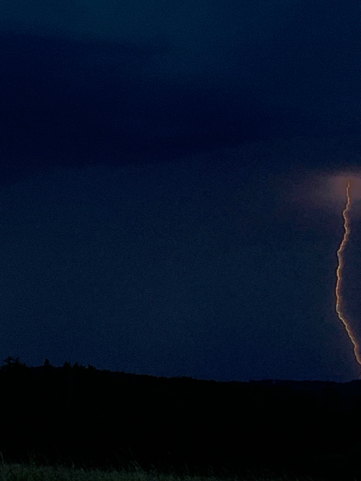 The image captures a dramatic natural phenomenon: a lightning strike illuminating the dark sky over a silhouetted landscape. The scene is set at dusk or night, with a deep blue-black sky dominating the upper portion of the frame. A jagged bolt of lightning, glowing with a bright, fiery orange-yellow hue, streaks vertically down from the upper right, appearing to descend towards the horizon. The lightning is the only source of intense light in the upper part of the image, creating stark contrast against the dark clouds.
Below the sky is a dark, undulating silhouette of a tree line or rolling hills. This feature is rendered almost completely black, making it difficult to discern specific details, though it suggests a rural or semi-rural environment. In the foreground, a dark band represents the ground, with hints of very faint green or brown texture that could be grass or low vegetation, suggesting a field or open area.
The image does not contain any people, structures, or man-made objects. The focus is entirely on the atmospheric event and the dark, natural surroundings. The location is specified as Oberquembach, Germany, a rural municipality in Bavaria, which aligns with the depicted landscape. The image presents a raw and powerful moment of nature's display.