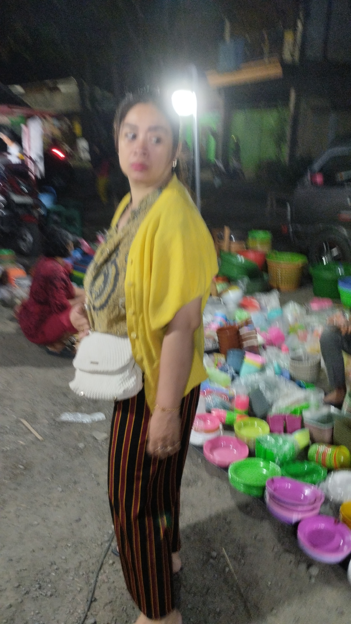 The image shows a woman standing in what appears to be a market in Sambungrejo, Indonesia. She is wearing a yellow cardigan over a patterned shirt and striped pants. She is carrying a white crocheted purse. The background is cluttered with various items, including plastic bowls and baskets in a range of colors. A light source, possibly a streetlamp, illuminates the scene. There are other people and vehicles visible in the background, suggesting a busy market environment. The ground is covered in gravel and dirt.
