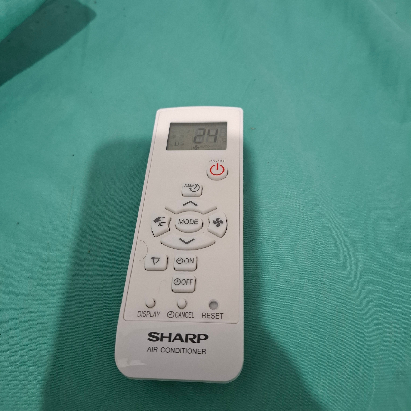The image shows a white remote control for a SHARP AIR CONDITIONER, lying on a teal-colored fabric surface. The remote control has a digital display showing "24" and "AUTO" to the left of a circular red power button with "ON/OFF" text. Below the power button are several other buttons arranged in a somewhat circular pattern. These include a "SLEEP" button with a crescent moon icon, a fan icon, a "MODE" button, and directional arrows. Further down, there are buttons labeled "JET", "ON", and "OFF", along with smaller buttons for "DISPLAY", "CANCEL", and "RESET". The SHARP logo and "AIR CONDITIONER" text are printed on the right side of the remote. The fabric surface appears to be slightly textured, with some subtle shadows and specks visible, suggesting it might be a bedsheet or a similar textile. The lighting is somewhat diffused, and there are no people or a specific time of day or weather indicated. The context of Reudeuep, Indonesia, suggests the remote is likely in a residential or commercial setting within that location. The overall scene is a still life of an electronic device.