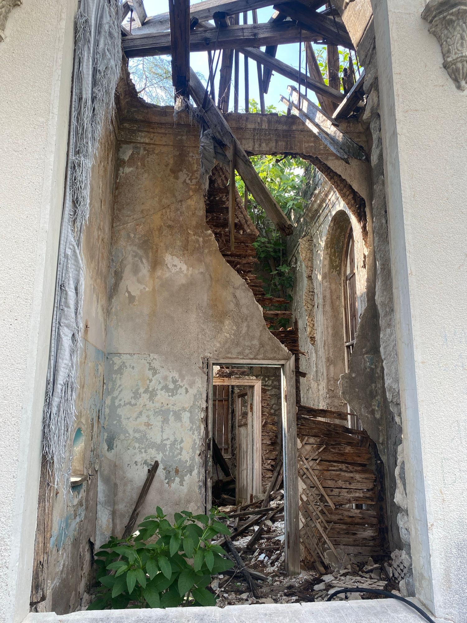 The image depicts the interior of a heavily damaged, possibly abandoned building in Psakhara, Abkhazia. The structure shows signs of significant decay and collapse, with a ruined roof exposing the sky above, supported by a network of broken wooden beams. The walls are crumbling, revealing plaster and brickwork, and some sections are covered in peeling paint. A damaged staircase is visible, constructed from splintered wood and debris. There are multiple doorways, some leading into darker, unknown spaces. Green foliage is growing through the rubble and within the structure, indicating a lack of human activity and the encroachment of nature. The overall impression is one of severe disrepair and neglect.