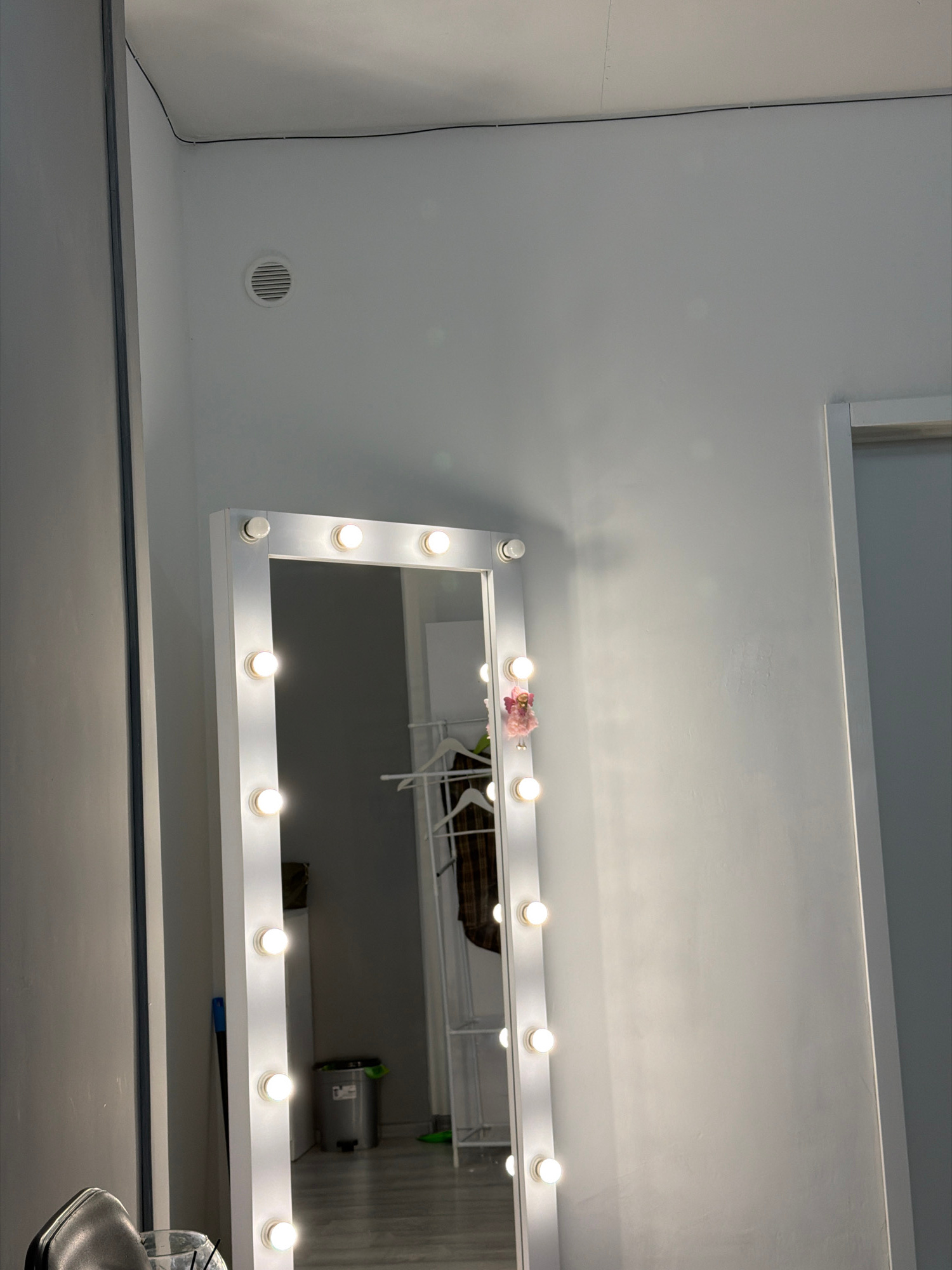 The image shows a full-length mirror with lights around its frame, positioned in a room. The room appears to be a dressing room or a makeup studio. The mirror reflects a clothing rack with garments, a trash can, and a broom. The walls are painted white, and there is a ventilation grate on the ceiling. The lighting is bright due to the illuminated mirror. The floor is a light-colored wood or laminate. The overall scene is clean and organized.