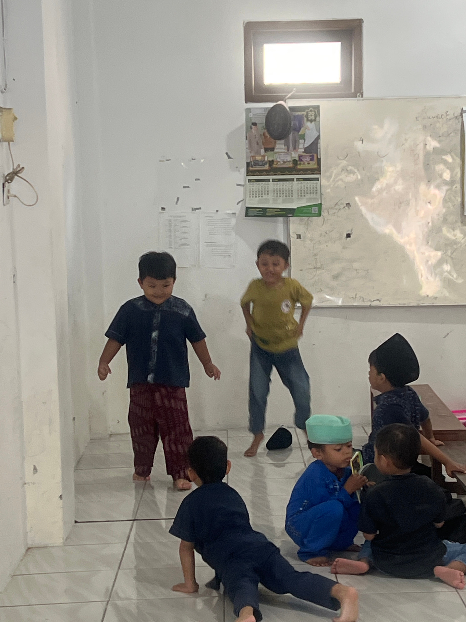 This image captures a scene inside what appears to be a classroom or community hall in Kecamatan Pati, Indonesia. The room is simple, with tiled floors and white walls. A window with a bright light is visible at the top of the frame, and a calendar hangs on the wall below it.
Several young boys are present, dressed in traditional Islamic clothing. Two boys stand in the center of the room, one in a dark blue shirt and patterned trousers, the other in a yellow shirt and blue jeans. Both appear to be engaged in some form of playful activity, with dynamic postures suggesting movement or laughter.
Other boys are seated or in kneeling positions on the floor. One boy in a blue outfit and a light green cap is interacting with another boy in a black shirt, possibly sharing a device. Another boy, wearing a black cap, is partially visible on the right, seated near a wooden bench. A fifth boy, dressed in a dark blue outfit, is positioned on the lower left of the frame, also appearing to be on the floor. The overall atmosphere suggests a casual and engaged group of children.