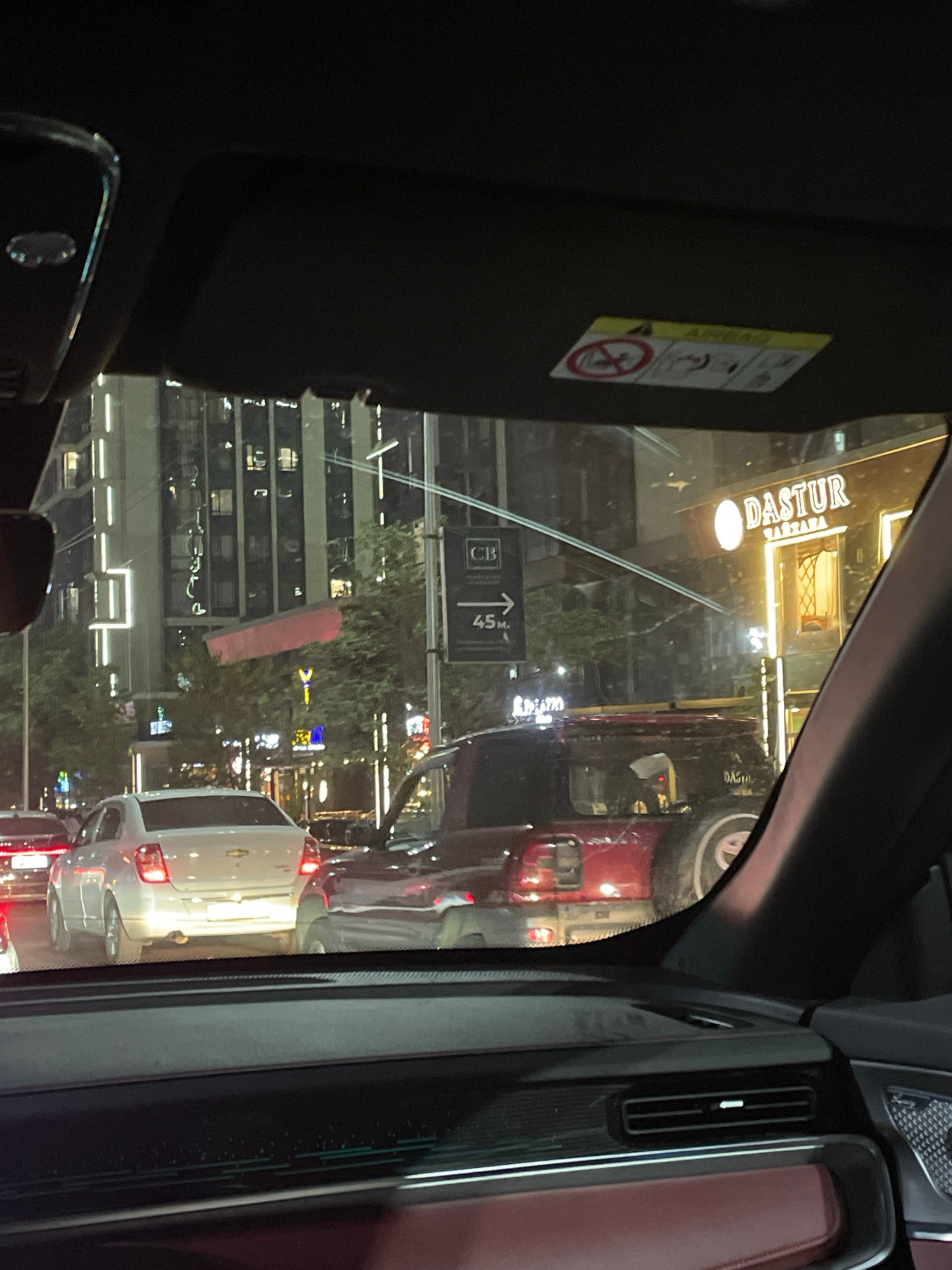 The image shows a view from inside a car, looking out through the side window. The scene is a city street at night, with buildings and traffic visible. The image is oriented vertically.
In the foreground, on the left side of the image, is the interior of the car. A black sun visor is visible, with a yellow and white sticker attached. The sticker contains several symbols and text, including a warning symbol with a person and a circle with a diagonal line through it, and the word "AIRBAG" is partially visible.
Through the window, the street is illuminated by streetlights and the lights from buildings and vehicles. Several cars are visible, including a red vehicle in the middle ground and a white vehicle in the right foreground. Buildings with illuminated windows line the street. There is a sign visible that reads "DASTUR". Another sign shows "45m".
The time of day is clearly night, indicated by the artificial lighting and the darkness of the sky. The weather appears to be clear. The location is identified as Almaty, Kazakhstan, though there are no specific visual cues in the image that directly confirm this without external information. The general atmosphere suggests an urban environment at night, possibly in traffic.