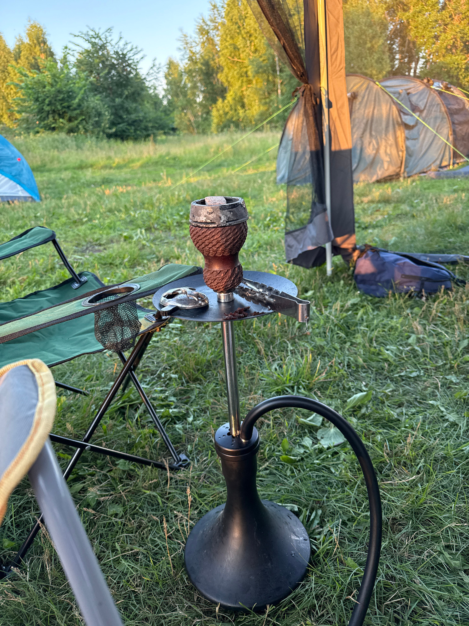 This image captures a camping scene in a grassy, outdoor setting, likely during the late afternoon or early evening due to the warm, soft lighting. The primary subjects are camping equipment and a hookah. In the foreground, a green and black foldable camping chair is prominent. Perched on a metal pole extending from behind the chair is a hookah pipe, with a textured brown bowl and a black hose. The hookah is angled, suggesting it might be in use or set up for a session.
Behind the chair and hookah, several tents are visible. One tent, partially unrolled and made of a translucent, greenish material, appears to be a tunnel tent or an inner tent. Another tent, or perhaps a tarp, in a dark grey or black, is also present. A blue backpack rests on the grass in the middle ground, adding to the camping atmosphere.
The background shows a line of trees and foliage, indicating a natural environment. The grass is green and interspersed with weeds and some dry blades, typical of a summer or late summer landscape. The sky is a pale, clear blue, hinting at pleasant weather conditions. The overall mood is relaxed and leisurely, consistent with a camping trip or outdoor gathering. There is no visible text in the image.