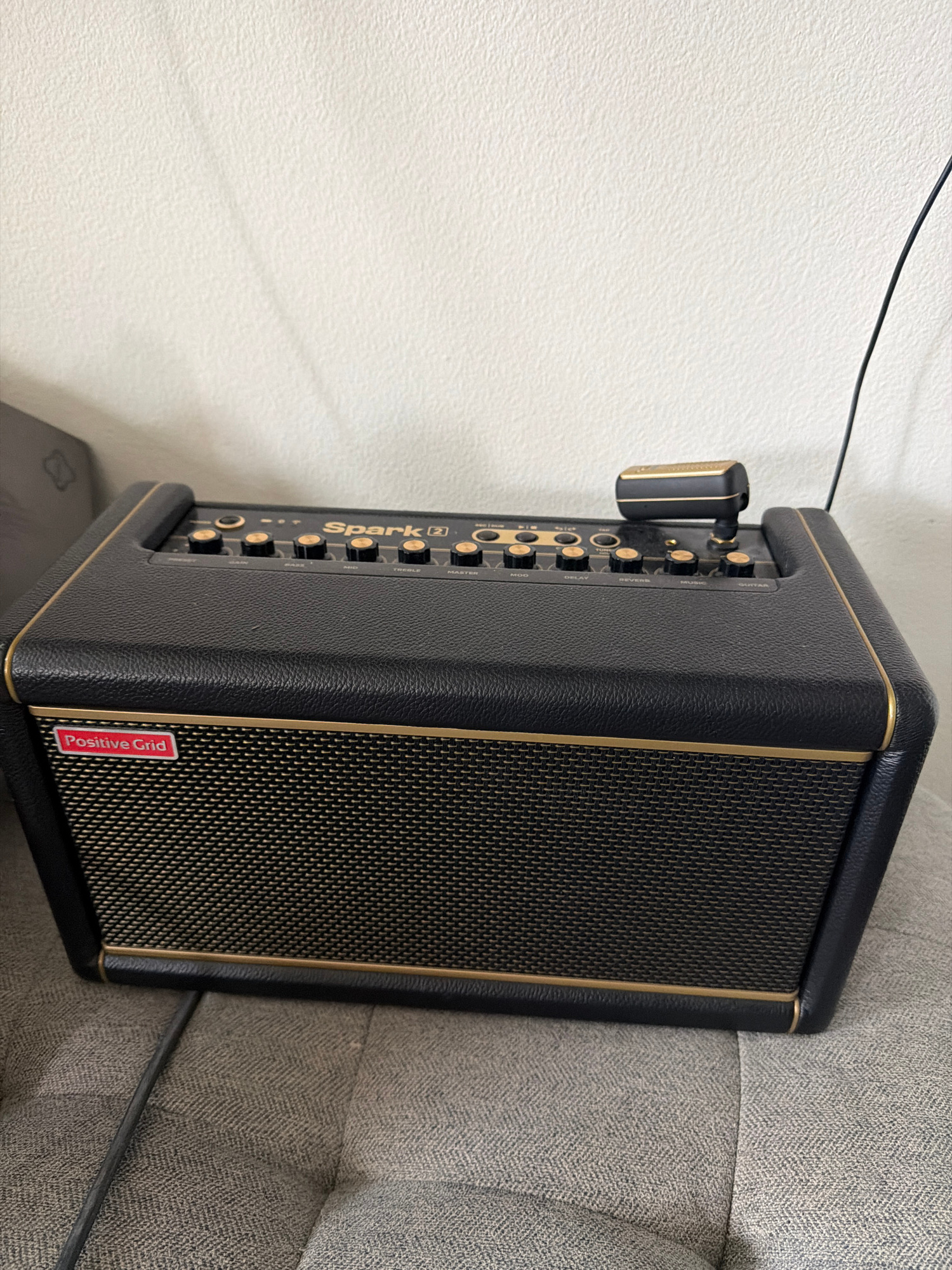 A black Positive Grid Spark 2 amplifier sits on a grey fabric surface, possibly a couch or chair, against a textured off-white wall. The amplifier has a black textured vinyl covering with gold piping around the edges. The front grill is also black with a gold trim, and a red and white "Positive Grid" logo is visible on the lower left. The top of the amplifier features several gold knobs and buttons, labeled with controls such as "GAIN," "BASS," "MID," "TREBLE," "MASTER," "MOD," "DELAY," "REVERBS," "MUSIC," and "GUITAR." A small black and gold microphone is attached to the top right of the amplifier. A black cable runs from the amplifier towards the bottom left of the frame, and another black cable, presumably for power, extends upwards towards the top right. The lighting suggests an indoor setting, possibly a home or studio. The location is described as Irvine, United States of America.