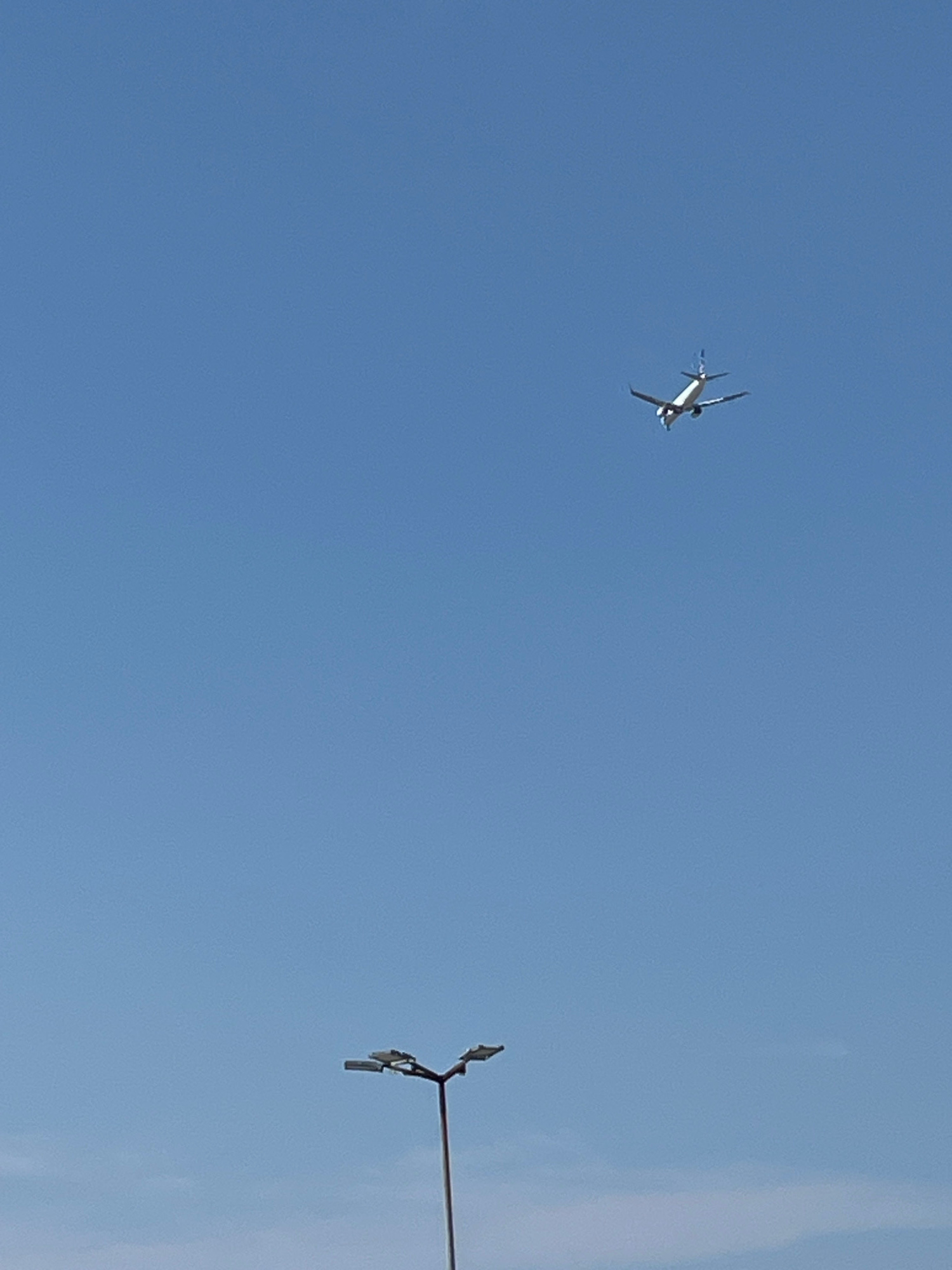 An airplane is in flight against a clear blue sky. In the foreground, a street lamp with two lights is visible. The image appears to be taken in Piaseczno, Poland.