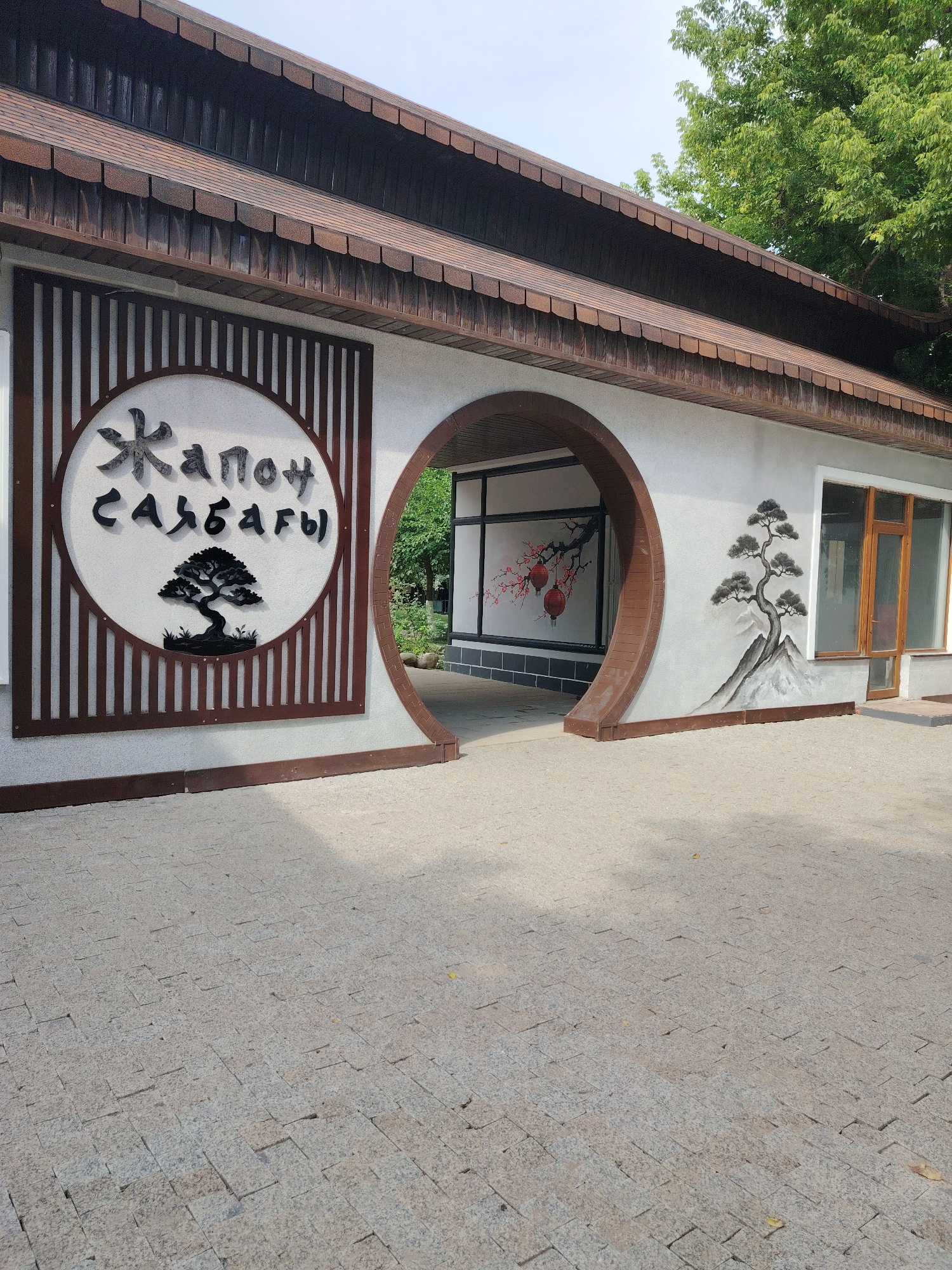 A modern building with Japanese-inspired architectural elements stands under a clear sky, partially obscured by lush green trees. The building features a dark, textured roof with a scalloped edge and a white facade. A prominent circular emblem on the left side displays stylized black lettering and a silhouette of a bonsai tree. To the right of the emblem, a rounded archway leads into a shadowed interior, where a mural of a blossoming cherry branch with red lanterns is visible. On the exterior wall to the right of the archway, a painted scene depicts a gnarled pine tree with dark branches and needles, set against a backdrop of gray mountains. The building has large windows with wooden frames. The ground in front of the building is paved with light-colored rectangular stones, casting dappled shadows from an unseen light source. The scene is static and devoid of human presence.