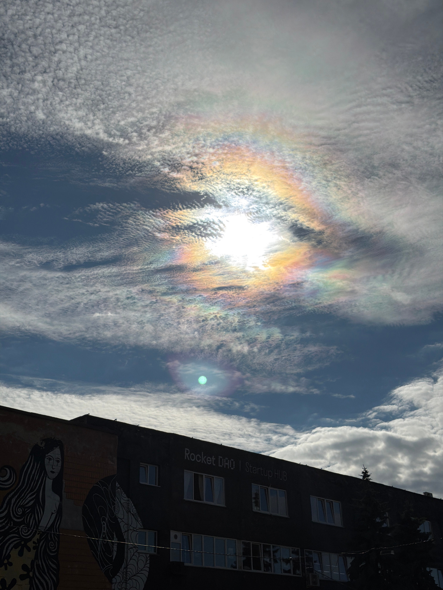 The image captures a rare atmospheric phenomenon, a cloud iridescence, occurring in the sky above a building in Minsk, Belarus. The iridescent clouds, exhibiting a spectrum of pastel colors, are situated above the sun, which is partially obscured. Below the sky, a dark-colored building spans the width of the frame. The building's facade features a mural on the left side, depicting a stylized female figure with long, flowing hair. Text on the building, legible as "Rocket DAO | Startup HUB," is visible in the upper section. The scene is a daytime shot, with a mix of blue sky and clouds, and the building appears to be an urban structure.