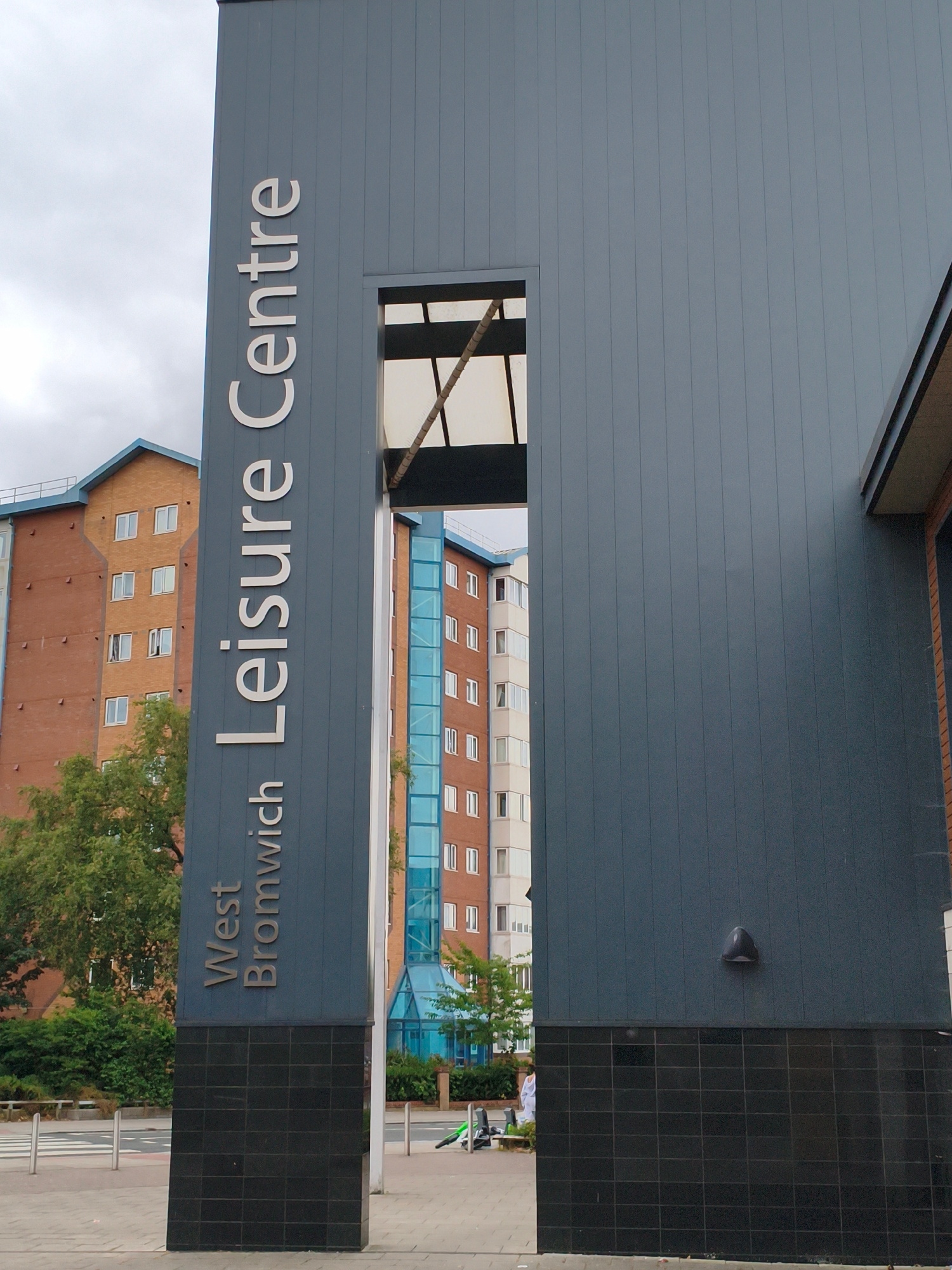 The image shows a vertical sign on the side of a building that reads "West Bromwich Leisure Centre." The sign is made of metal letters on a dark blue panel. In the background, there are multi-story brick buildings with many windows. The sky is overcast. The foreground shows a paved area with bollards and a pedestrian crossing. A person in white clothing is visible in the background walking towards the building. The overall scene suggests an urban environment.