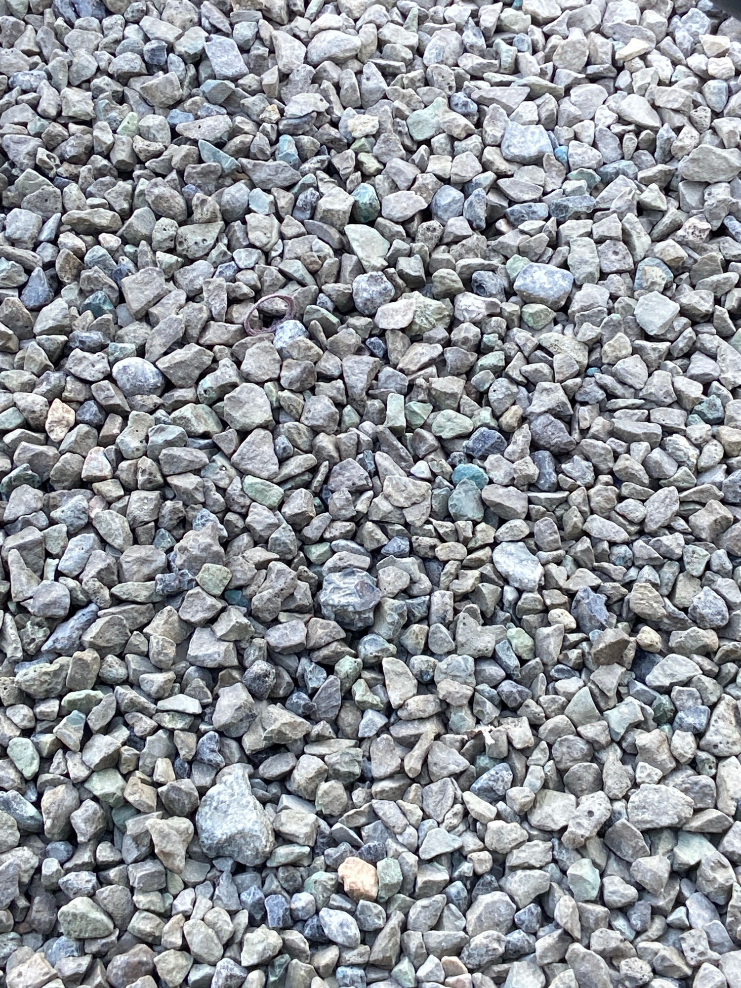 A close-up, overhead shot displays a dense, textured surface of small, angular, greyish-blue and light brown gravel. The stones vary slightly in size and shape, with some appearing more weathered and rounded than others. A thin, reddish-brown ring, possibly a piece of wire or plastic, is visible near the top center of the frame. The lighting suggests an outdoor environment, though no specific landmarks or features indicating Chelyabinsk, Russia are present. The image appears to be a still photograph with no apparent action or interaction occurring.