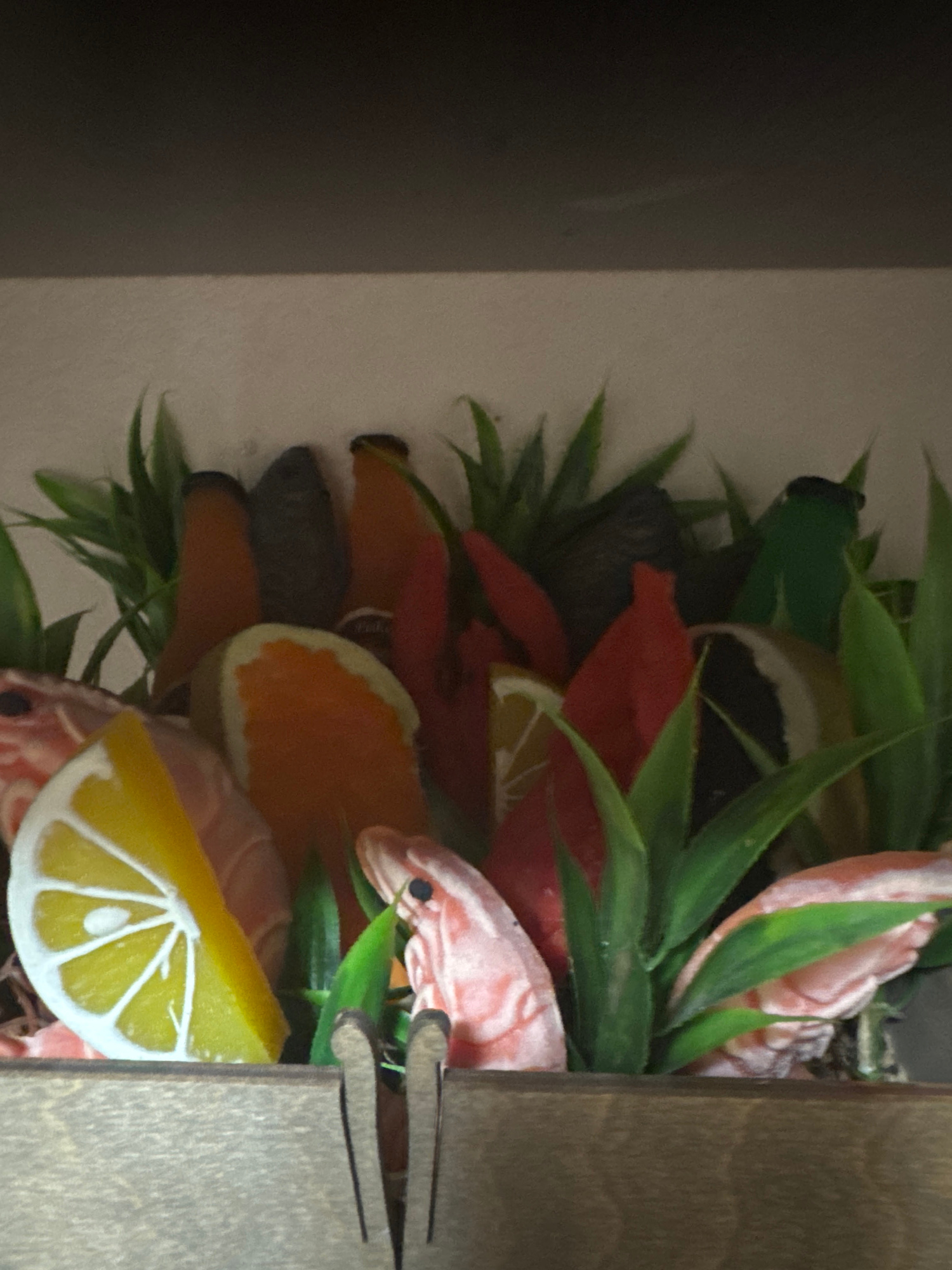 This media file is a close-up, vertically oriented photograph showcasing a decorative arrangement of artificial fruits and shrimp. The primary subjects are several slices of citrus fruits, including oranges and lemons, and pinkish shrimp, interspersed with green, leafy artificial foliage. The arrangement appears to be positioned within a wooden container or shelf, with the texture of the wood visible on the right side of the image. The overall impression is of a still-life display, possibly for decoration or as part of a larger artistic composition. There are no people or animals visible in the image. The lighting suggests an indoor setting with moderate illumination, casting soft shadows. No text is visible. The location context provided is "Змейка, Russia," which offers geographical information but is not directly discernible from the visual content of the image itself.