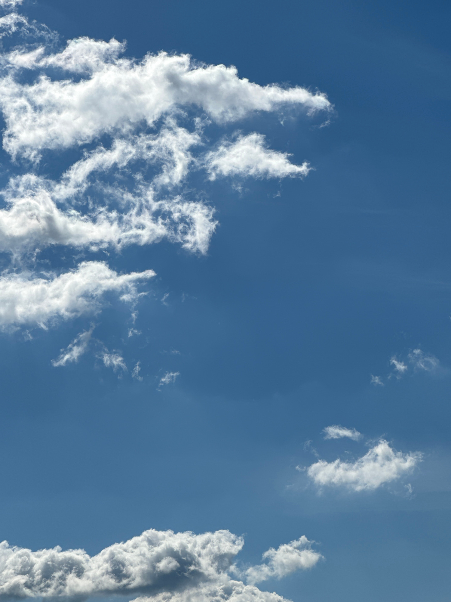 The image shows a bright blue sky with various white clouds. In the upper left quadrant, there are several large, fluffy clouds with some wispy formations. Towards the bottom of the image, a thicker band of clouds runs horizontally across the frame, with some darker, shadowed areas suggesting depth. Scattered throughout the sky are smaller, more isolated clouds. The overall impression is of a clear day with scattered cloud cover. There are no people, structures, or other identifiable objects visible in the image.