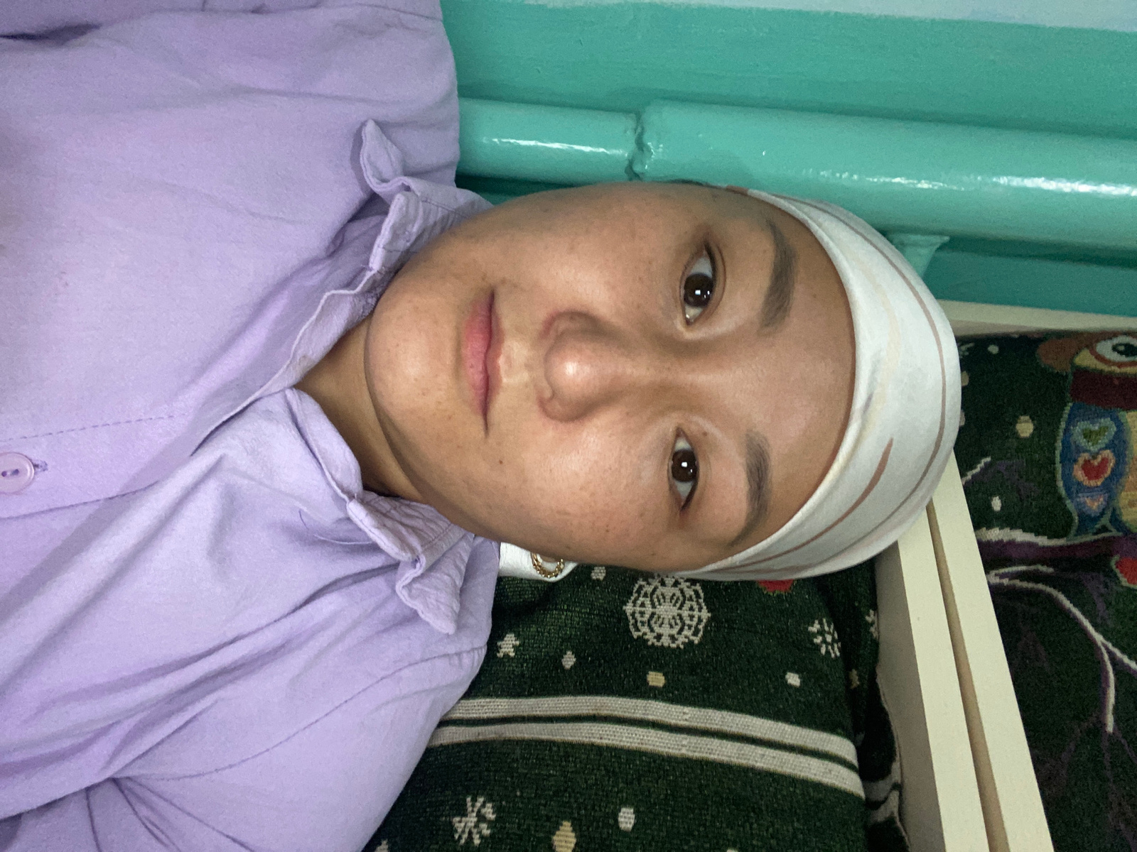 A medium shot photograph, taken from a slightly elevated angle and oriented to the left, shows a young woman lying down. She is wearing a lavender collared shirt and a white headscarf with thin brown stripes. Her head is resting on a dark green patterned blanket. The background features a turquoise colored wall with pipes running horizontally across it. The room appears to be a bedroom or a similar domestic space. The woman's expression is neutral. The image is dimly lit, with a slight shadow cast on her face from the left side.