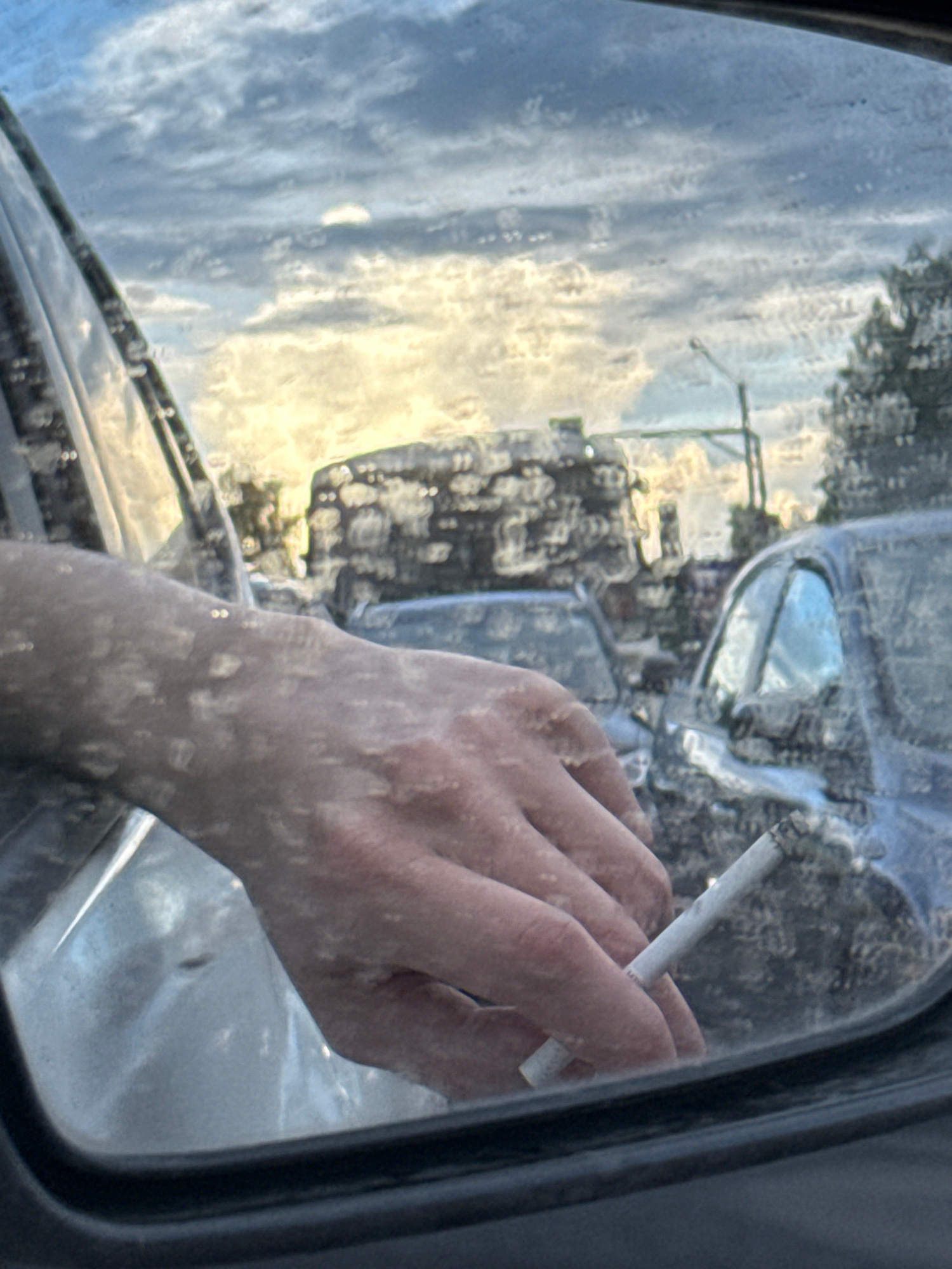 The image captures a hand holding a lit cigarette, viewed through a car window. The hand, appearing to be an adult's, is positioned on the right side of the frame, its fingers wrapped around the cigarette. The cigarette is burning, with visible ash at the tip.
The scene outside the car window is a reflection, showing a cloudy sky with streaks of yellow light suggesting either dawn or dusk. The reflection also reveals trees and the indistinct shapes of buildings or vehicles. The overall impression is that the car is either parked or moving slowly, and the perspective is from inside the vehicle, looking outwards.
There are water droplets on the car window, creating a slightly distorted and hazy view of the outside. The lighting is diffused, with the bright sky contrasting with the darker interior of the car. The mood is somewhat contemplative, possibly indicative of a moment of pause or reflection while traveling.
The location context of "Агрогородок, Russia" provides a geographical setting, suggesting a rural or semi-rural environment. However, there are no specific visual cues within the image that definitively confirm this location. No visible text is present in the image.