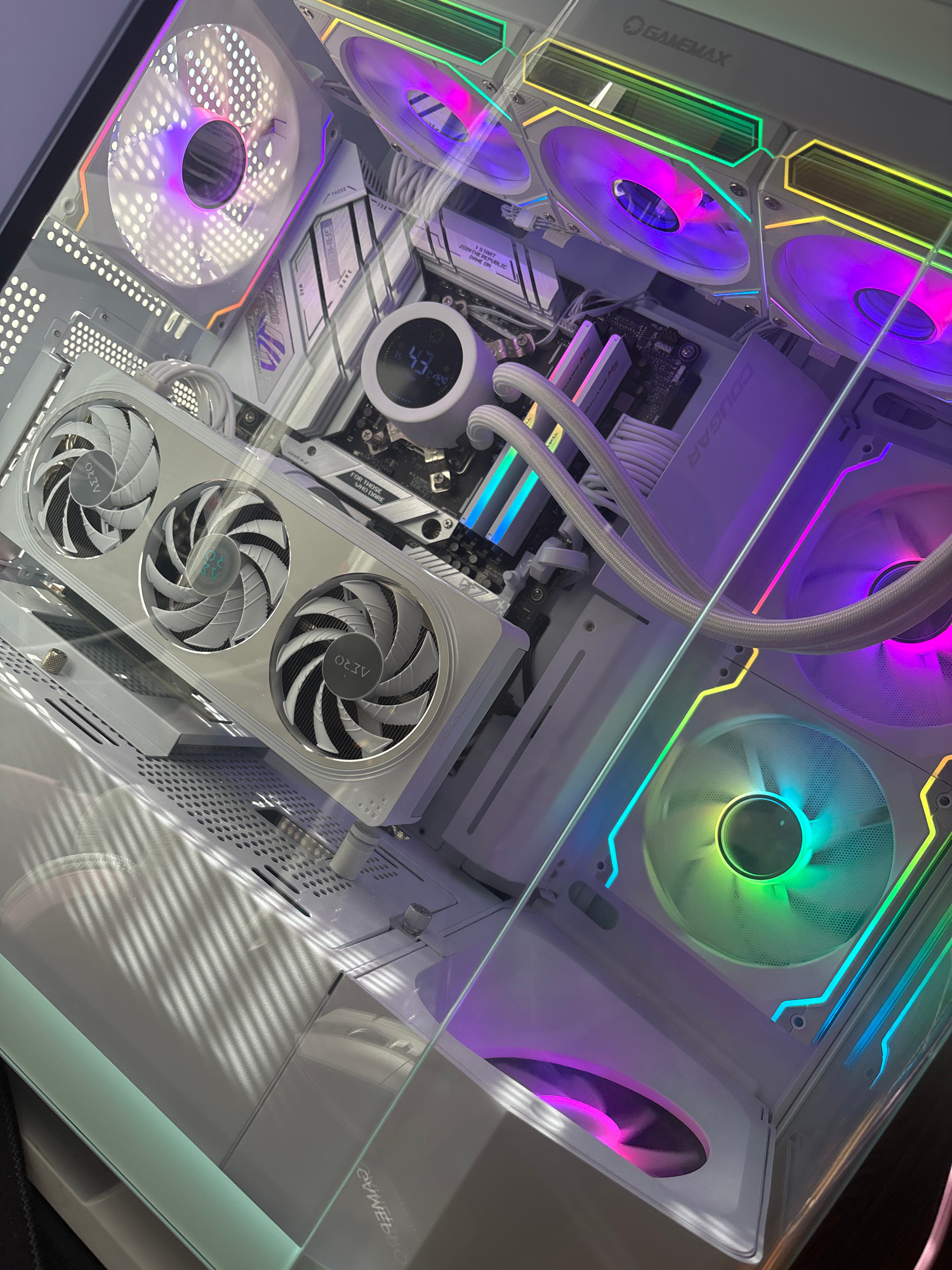 The image shows a white computer tower with multiple RGB fans. A graphics card with three fans is mounted vertically. There is a liquid cooler with a circular display on the motherboard. The computer case has a clear side panel, allowing a view of the components inside. The lighting is provided by the RGB fans, which illuminate the interior with various colors. The computer is placed on a dark surface. The image is taken from a high angle, looking down into the computer case.