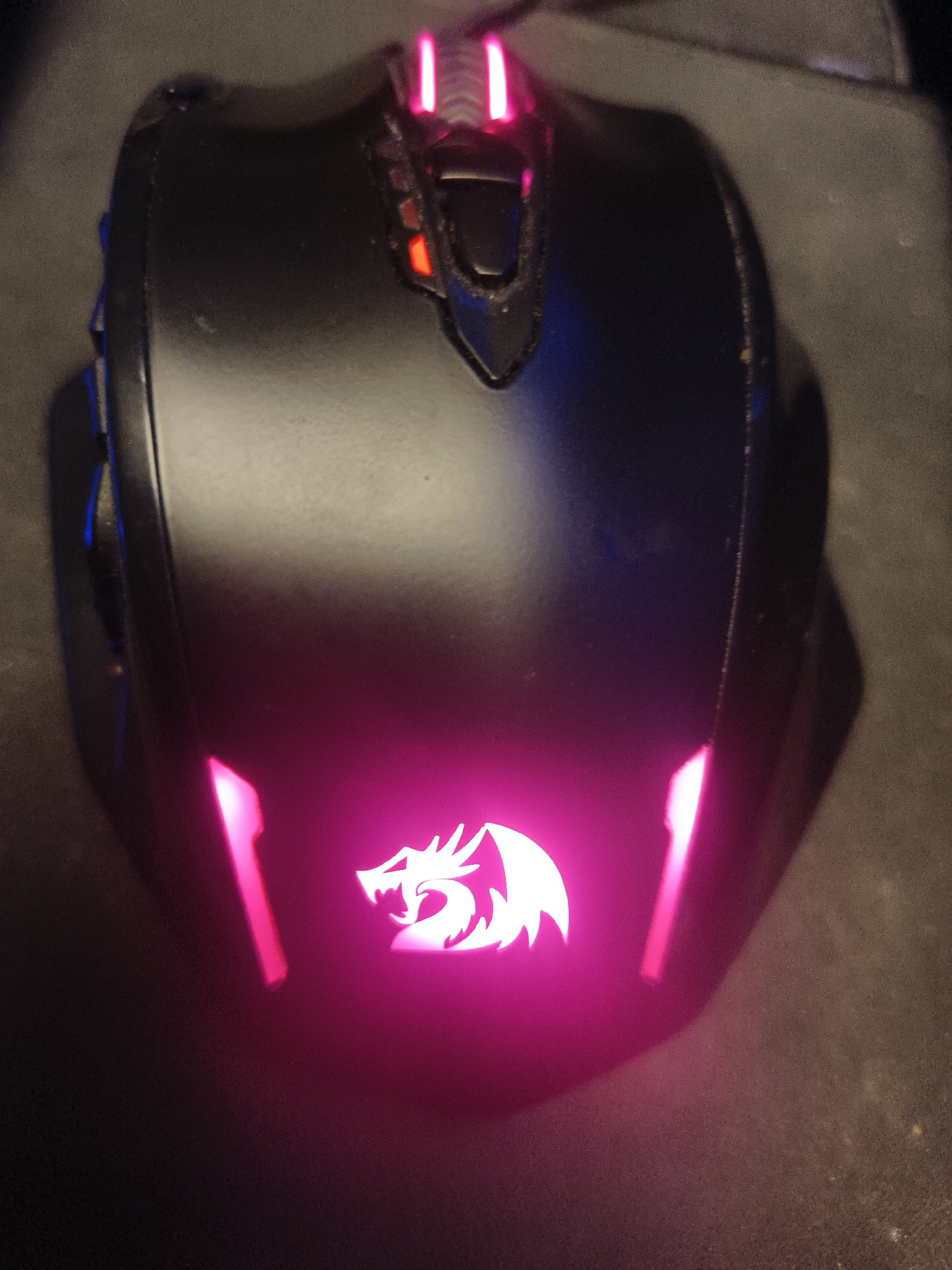 A black computer mouse with pink LED lighting sits on a dark gray surface. The mouse features a stylized dragon logo illuminated in white light against a pink glow. Two pink LED strips are visible at the top of the mouse, and two more pink LED strips are positioned on the lower front section. The mouse appears to be on a mousepad. There is no indication of the city of Kuta Selatan, Indonesia.