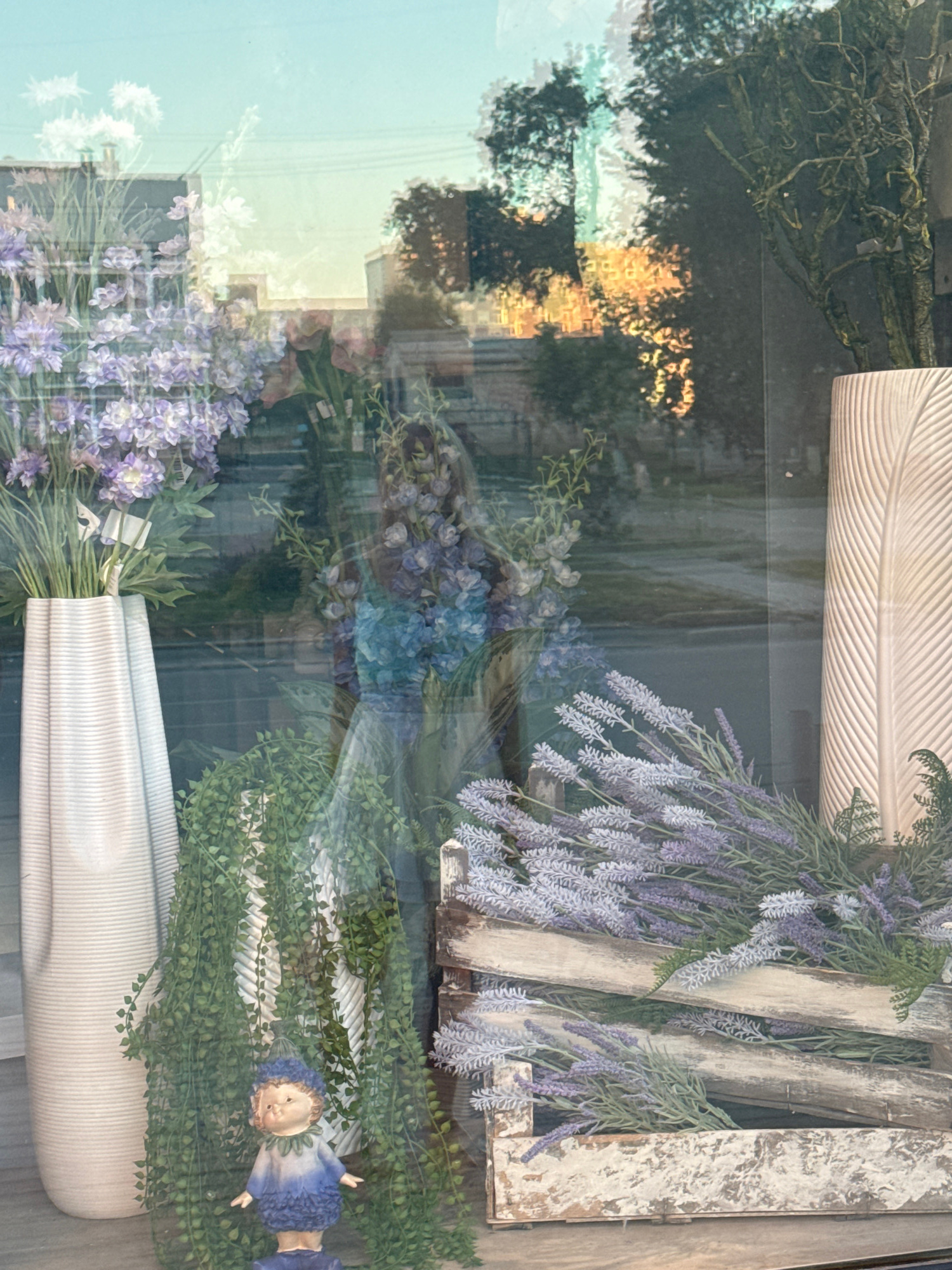 The image is a reflection in a storefront window in Barnaul, Russia, displaying a variety of artificial floral arrangements and decorative items. In the foreground, a white, ribbed vase holds a bouquet of purple and white flowers, with cascading green foliage to the left. A small, blue-clothed figurine stands beneath the foliage. To the right, a distressed wooden crate is filled with lavender sprigs. Behind these items, a tall, white vase with a feather-like pattern is visible. The reflection shows trees, buildings, and a sunset-lit sky, indicating an outdoor environment. The overall scene depicts a retail display, likely for a flower shop or home decor store.