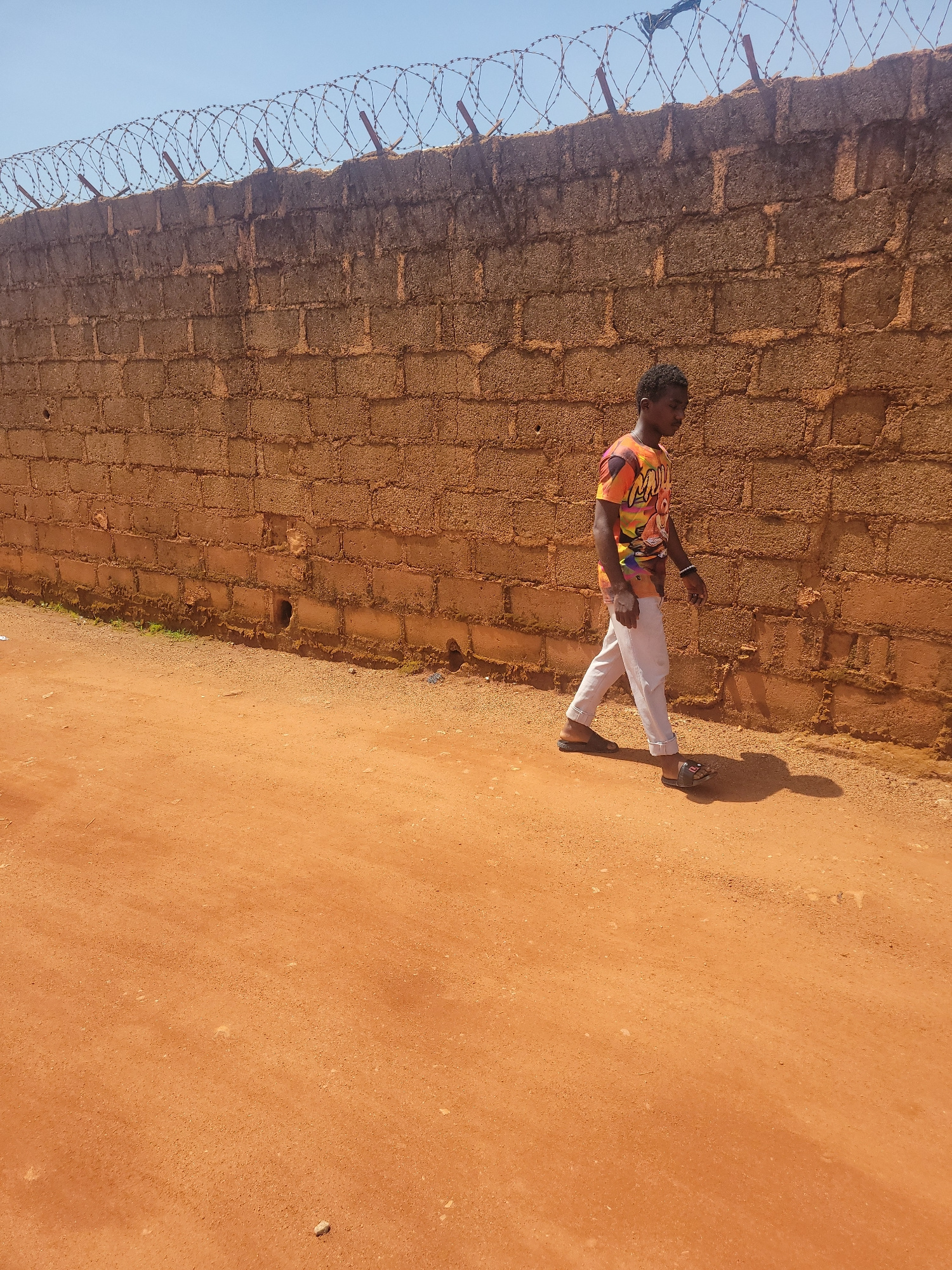 A young man walks along a dusty, orange-colored path next to a tall, rough brick wall topped with barbed wire. He is wearing a vibrantly colored t-shirt with an abstract design and light-colored trousers. He is also wearing dark sandals and has a bracelet on his left wrist. The sun is casting a shadow of the man on the path. The wall is made of roughly textured, rectangular bricks and appears to be a boundary for a property. The sky is clear and blue. This scene is typical of many areas in Jos, Nigeria, where such walls and dusty paths are common.
