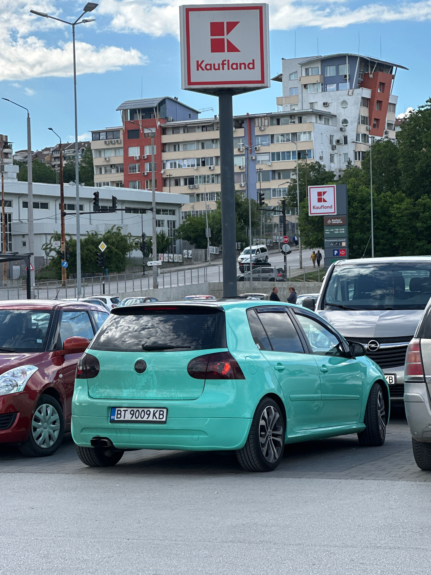 A teal Volkswagen Golf hatchback with license plate BT 9009 KB is parked in what appears to be a parking lot in Veliko Tarnovo, Bulgaria. The car is facing away from the camera, and its rear bumper, taillights, and alloy wheels are visible. To the left of the Golf, a red Suzuki Swift is parked. In the background, several modern buildings with mixed architectural styles can be seen, some featuring colorful facades and balconies. A large red and white sign for "Kaufland" is prominently displayed on a pole in the foreground. Additional traffic lights and street signs are visible in the midground, suggesting an urban or commercial area. The sky is partly cloudy.