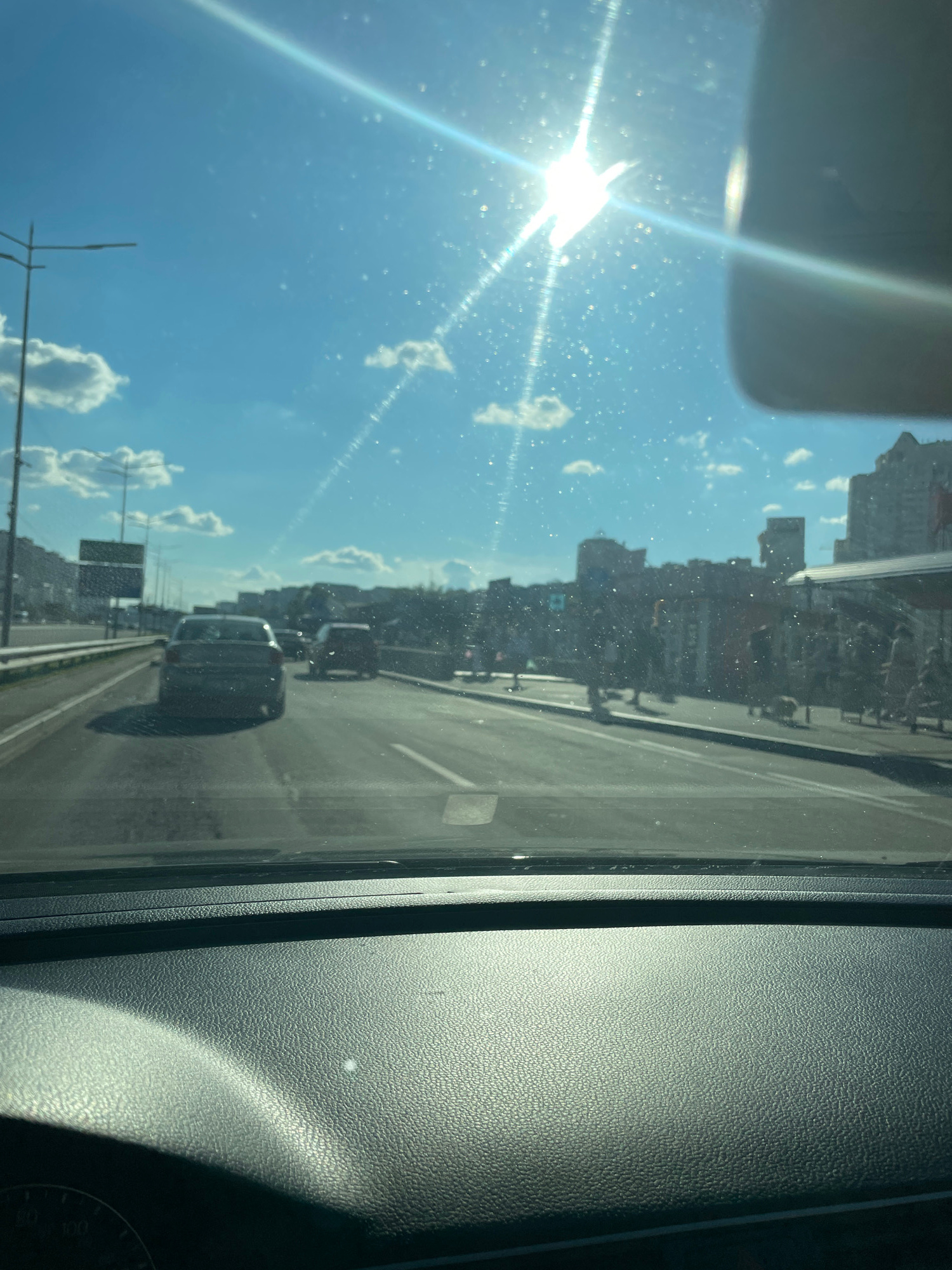 The image is taken from inside a vehicle looking forward through the windshield on a sunny day. The sky is blue with scattered white clouds. The sun is bright and high in the sky, causing glare and lens flare. There are specks of dust or debris on the windshield.
The vehicle is on a multi-lane road, likely a highway or a major thoroughfare, in Kyiv, Ukraine. Several cars are visible on the road ahead, moving in the same direction. The road is paved and appears to be in good condition. Lane markings are visible.
On the right side of the road, there is a sidewalk or pedestrian area with people walking. Buildings are visible in the background, appearing to be a mix of modern and older architecture, consistent with an urban environment. Some of these buildings are tall, suggesting a city center or a densely populated area. Streetlights are visible along the side of the road.
On the left side of the road, there are more lanes of traffic and what appears to be a highway overpass or a large sign structure.
The interior of the vehicle is partially visible in the foreground, showing the dashboard. The dashboard is black and textured.