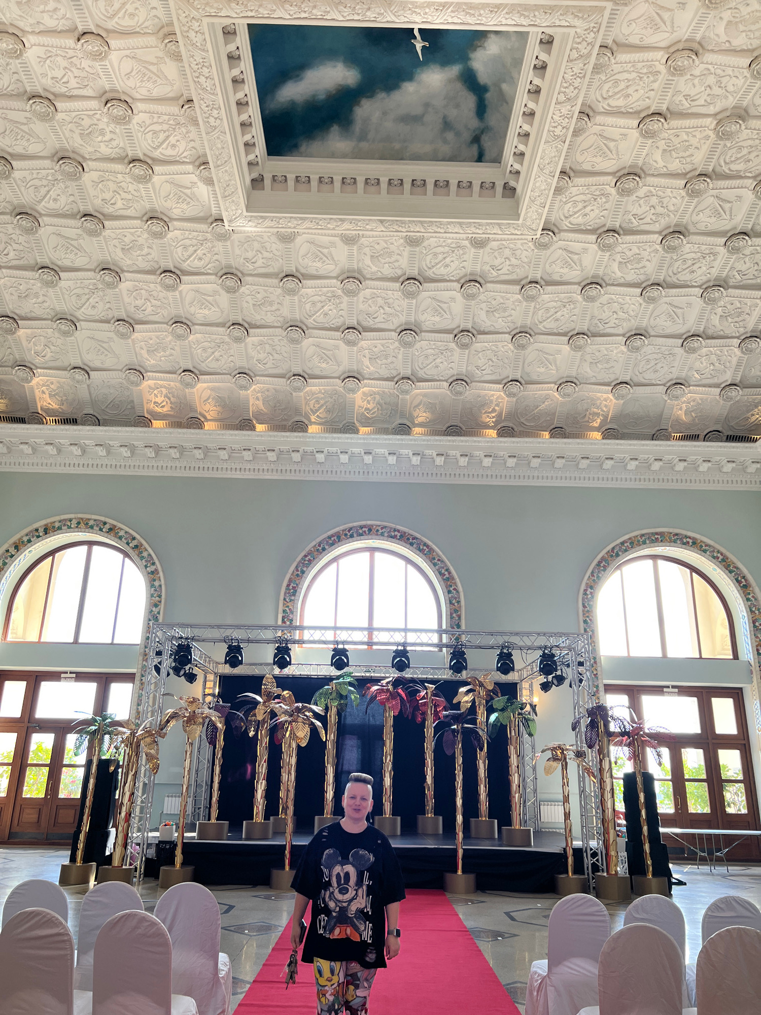 The image captures an interior scene with a high, ornate ceiling featuring a repetitive pattern of embossed rosettes and heraldic crests. A large framed artwork depicting a bird flying through clouds is visible on the ceiling. To the right, the scene transitions to a space with light blue walls and arched windows. A stage is set up with golden palm tree decorations and professional lighting equipment. In front of the stage, a red carpet leads to rows of white-covered chairs, suggesting an event such as an award ceremony or a performance. A person, wearing a black t-shirt with a Mickey Mouse print and colorful patterned pants, stands on the right side of the frame, holding a small bouquet of flowers. The person appears to be smiling, indicating a positive or celebratory atmosphere. The lighting suggests it is daytime, with natural light filtering through the windows. The location is identified as Sochi, Russia.