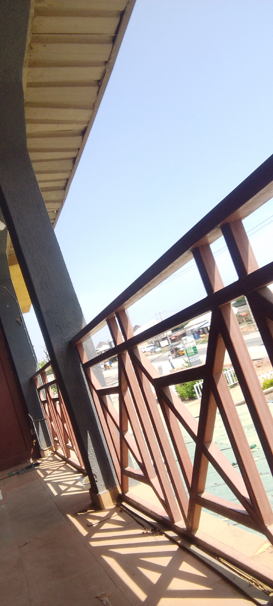 The image shows a view from a balcony or elevated walkway, looking outwards towards a street scene in Suleja, Nigeria. The foreground is dominated by a reddish-brown metal railing with a geometric design, casting distinct shadows on the tiled floor below. To the left, a textured, dark gray pillar supports an overhead structure with light-colored wooden planks. Beyond the railing, the street is visible, with several buildings, some with corrugated iron roofs. Various vehicles, including cars and what appears to be a motorized tricycle (Keke Napep), are present on the street. A few pedestrians can be seen, one standing near a street sign. The sky is clear and bright blue, indicating daytime. The overall scene depicts a typical urban environment in Suleja.