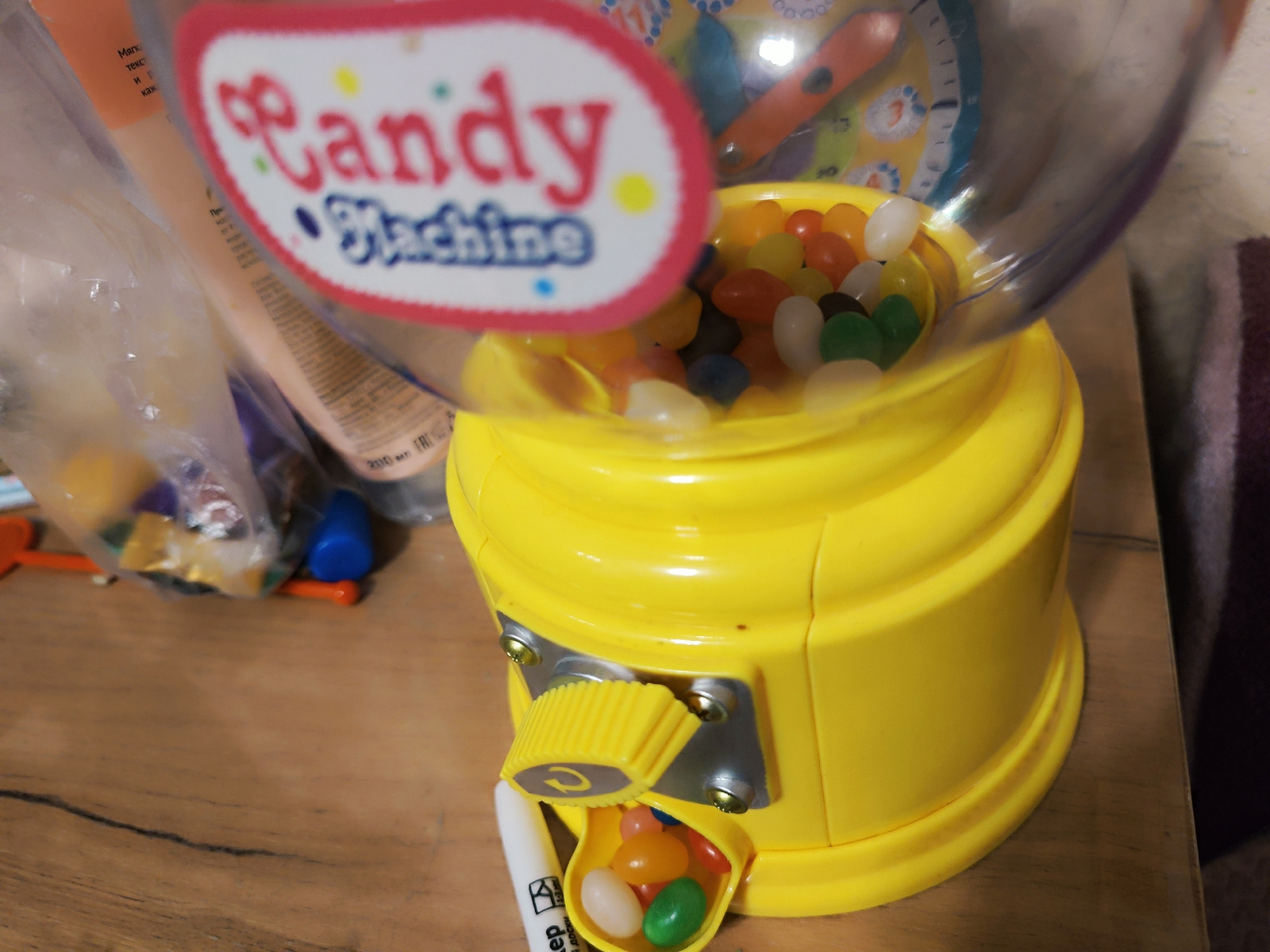 A bright yellow, plastic candy machine with a clear plastic dome filled with colorful jelly beans sits on a wooden surface. The dome has a rounded, oval label with red trim that reads "Candy Machine" in pink and blue lettering. A slot below the dome dispenses the jelly beans into a yellow catcher. A white marker with black text is positioned next to the catcher. In the background, various items are visible, including plastic bags containing more candy or toys, and part of a striped fabric. The scene appears to be indoors, possibly in a home setting. There is no indication of the city of Makiivka, Ukraine, in the image.