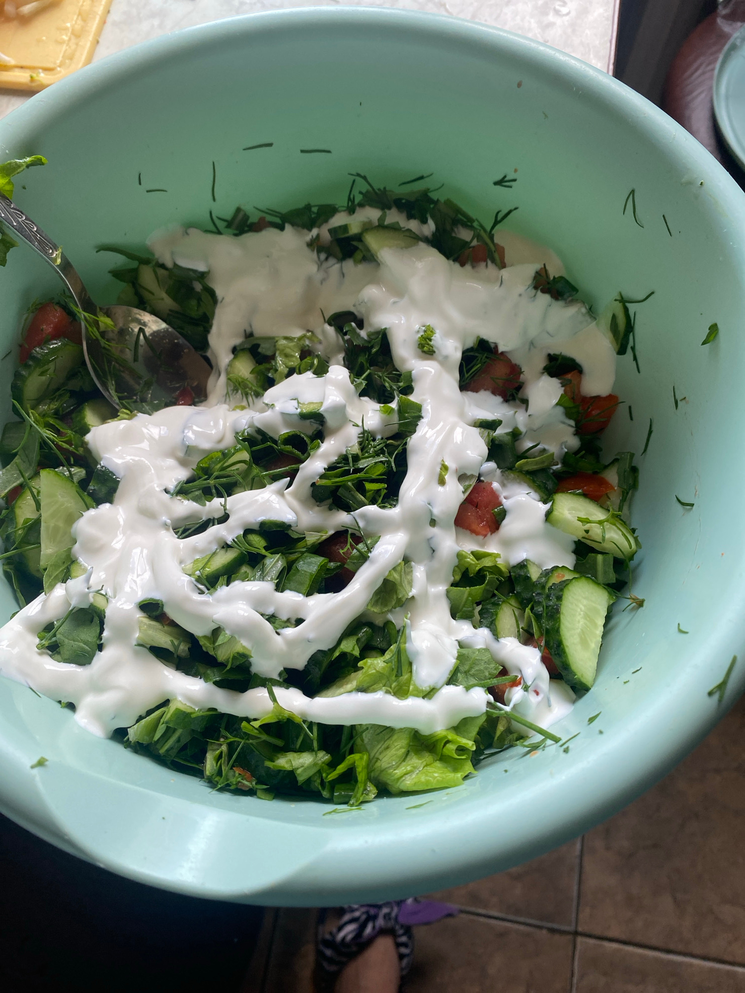 An overhead, medium shot captures a pale blue plastic bowl filled with a salad. The salad consists of chopped lettuce, cucumber slices, and diced tomatoes, all mixed with fresh herbs, possibly dill or parsley. A creamy white dressing, likely sour cream or yogurt-based, is drizzled generously over the top. A silver spoon is partially submerged in the salad on the left side of the bowl, suggesting it's being prepared or served. The bowl sits on a tiled floor, with a glimpse of what appears to be a light-colored countertop in the upper left corner. The lighting is natural and diffused, casting soft shadows. The image is devoid of any specific identifying landmarks or features that would pinpoint its location within Krupki, Belarus.
