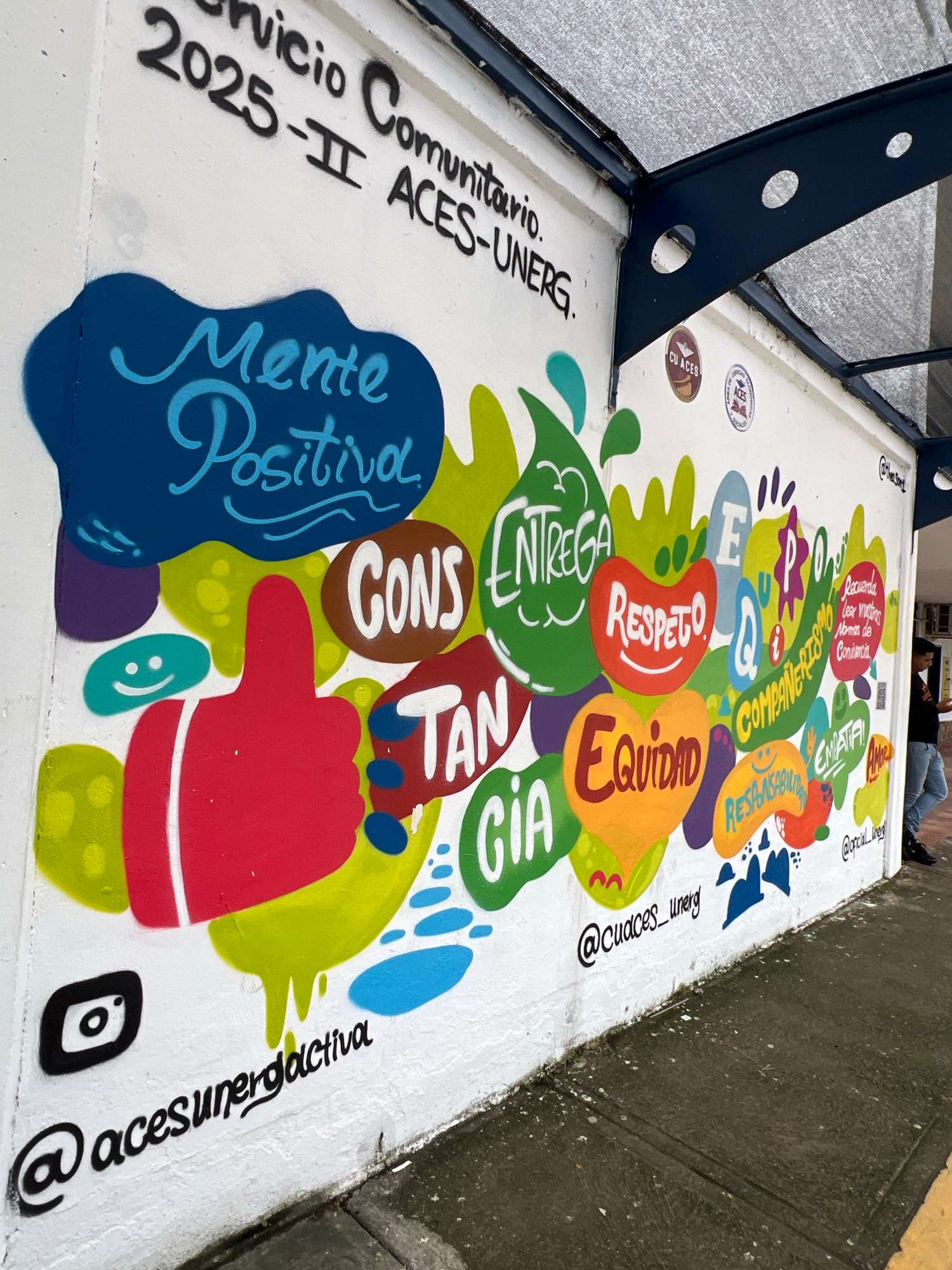 This media file captures a vibrant mural painted on a white surface, likely part of an urban installation or public art project in San Juan de Los Morros, Venezuela. The mural is characterized by colorful, abstract shapes and handwritten text in Spanish, promoting community values.
Prominent words and phrases include "Servicio Comunitario," "Mente Positiva," "Entrega," "Respeto," "Equidad," and "Amor," suggesting a theme of positive social contribution and shared values. The text "2025-II ACES-UNERG" likely refers to a specific program or initiative. Several social media handles, such as "@acesunerdeactiva," "@cuaces_unerg," and "@opcial_weg," are visible, indicating an online presence associated with the artwork.
The mural features a stylized red hand pointing, and a blue outline of a head with "Mente Positiva" written inside. Small decorative elements like smiley faces and blue splatters add to the playful and engaging aesthetic. The artwork appears to be located outdoors, adjacent to a grey, textured surface that could be a sidewalk or wall, and a dark blue metal structure is visible on the left, possibly part of a bench or railing.
The overall impression is one of positive community engagement and artistic expression within an urban setting. The sunlight suggests it is daytime, and the clear visibility of the artwork indicates fair weather. There is a person visible in the background, casually dressed and seated, suggesting a relaxed atmosphere.