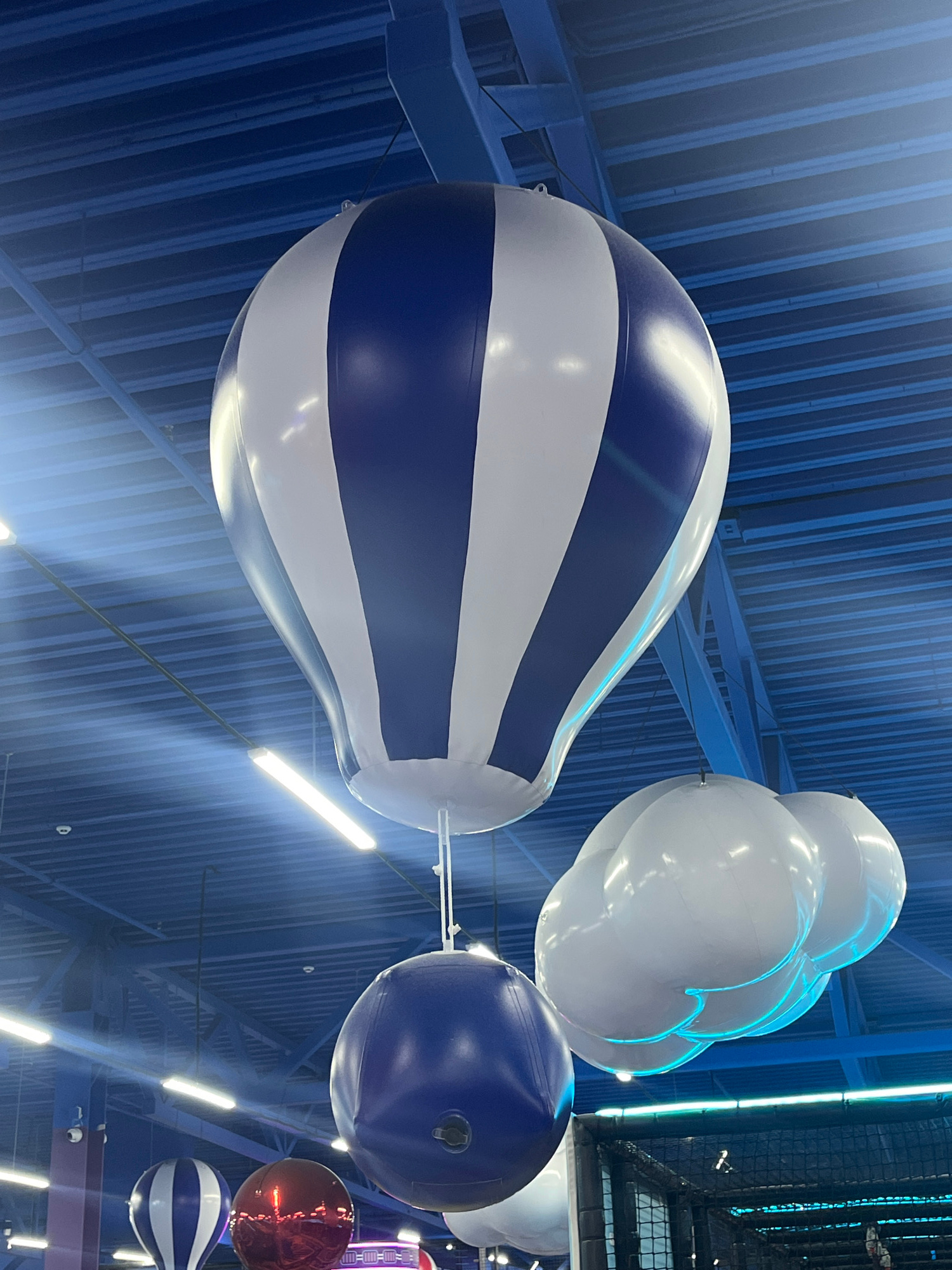 The image shows a large, inflatable hot air balloon with blue and white stripes suspended from a blue, ribbed ceiling. Below the balloon, a smaller blue inflatable ball hangs. To the right, a cluster of white inflatable clouds with blue lighting is also suspended. In the background, other inflatable objects, including a red ball and another striped balloon, are visible. The ceiling has fluorescent lights. The scene appears to be indoors, possibly in an entertainment venue. The location is the city of Kostanay, Kazakhstan. There are no people visible in the image.