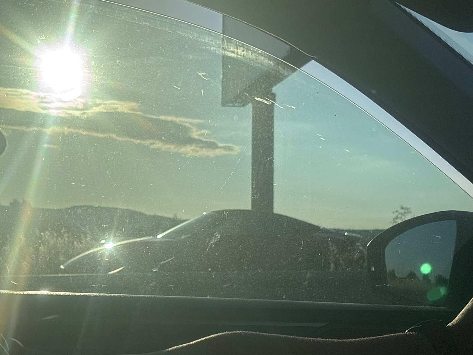The image captures a view from inside a car, looking out through the driver's side window. The sun is bright and high in the sky, suggesting it is midday. Lens flare from the sun creates streaks of light across the window and the scene. The window is dirty, with numerous smudges and streaks visible.
In the foreground, the dark interior of the car is visible, including a portion of the dashboard and the edge of the steering wheel. A hand is resting on the steering wheel, indicating the car is likely in motion or stopped but occupied by a driver.
Outside, the landscape is somewhat obscured by the glare and the dirty window. In the middle ground, a dark-colored car is visible, its shape suggesting a sleek, possibly sporty vehicle. Behind the car, rolling hills or mountains form the horizon line, covered in what appears to be dry vegetation.
A tall, dark structure, possibly a signpost or a utility pole, stands in the background, casting a shadow. The sky is a pale blue, with a few wispy clouds scattered around. The overall impression is a sunny day in a rural or semi-rural setting, possibly during a road trip. There is no visible text in the image. The location context provided is Aheloy, Bulgaria.