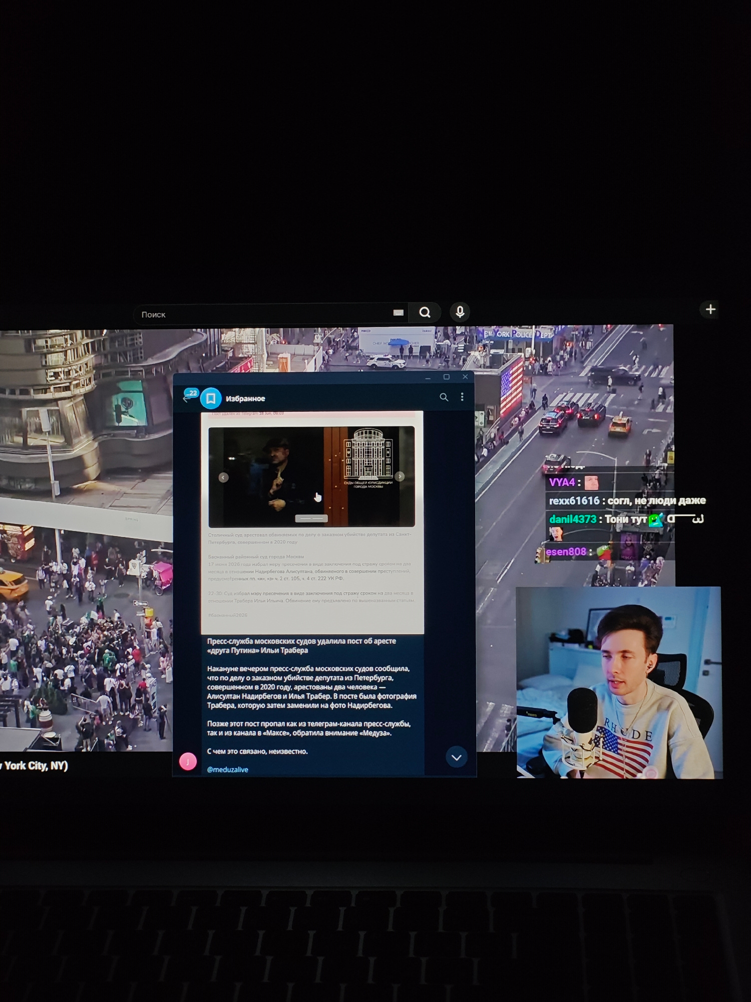 The image shows a computer screen displaying a live stream with a streamer in the bottom right corner. The stream itself shows a bird's-eye view of a busy city street, likely in New York City, given the text "New York City, NY" in the bottom left. In the center of the screen, a window is open with what appears to be a news article or social media post. The article discusses an arrest related to a murder case involving a deputy in St. Petersburg, Russia, and mentions the involvement of the press service of the Moscow courts. The article is in Russian, and contains text such as "Столичный суд арестовал обвиняемых по делу о заказном убийстве депутата из Петербурга" which translates to "The capital court arrested the accused in the case of the ordered murder of a deputy from St. Petersburg." The streamer is wearing a grey sweatshirt with an American flag design.