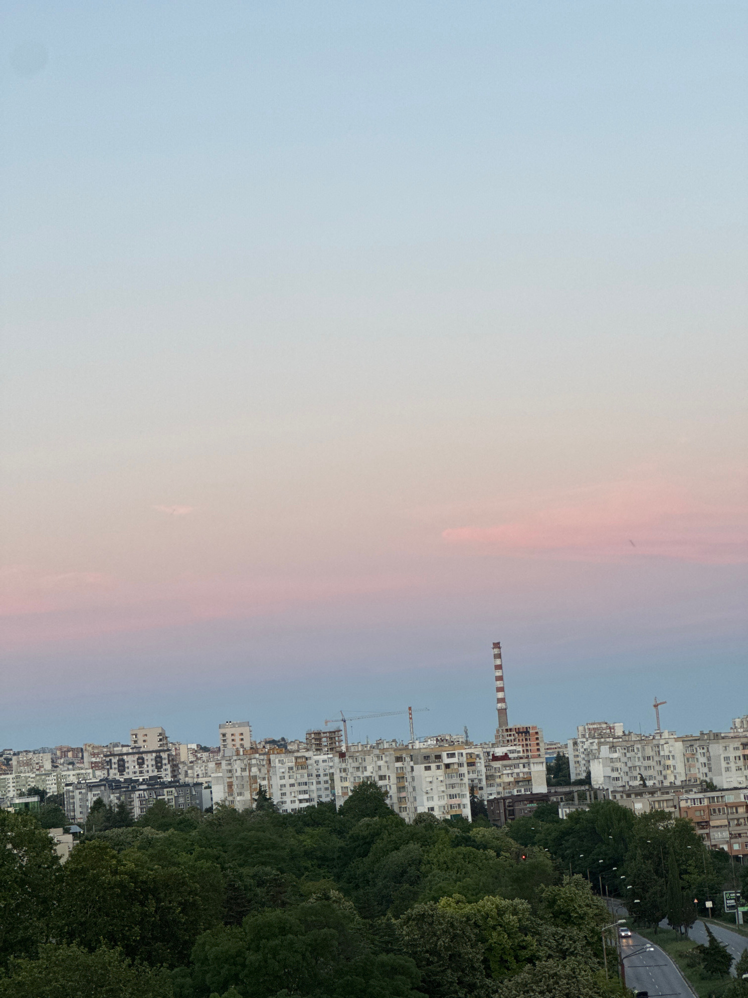 The image captures a panoramic view of Varna, Bulgaria, during what appears to be twilight. The sky exhibits a gradient of soft blues and pinks, indicative of sunset or sunrise. Dominating the mid-ground is a dense cluster of apartment buildings, characterized by their uniform, utilitarian architecture common in post-socialist urban development. Among these residential blocks, a tall, striped industrial chimney stands out, its red and white bands creating a stark contrast against the pastel sky. Construction cranes are visible, suggesting ongoing development in the city. In the foreground, a lush canopy of green trees partially obscures the lower parts of the buildings, creating a natural buffer between the urban landscape and the viewer. A winding road with streetlights and a few vehicles is visible on the right side, indicating a populated area. The overall scene conveys a sense of a bustling city transitioning into evening, with a blend of residential, industrial, and natural elements.