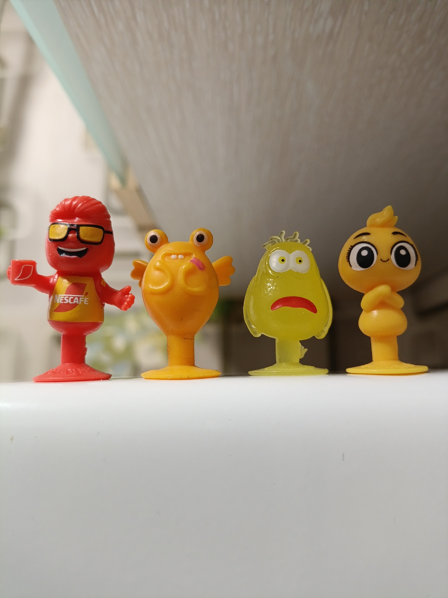 Four plastic figurines are displayed in a horizontal line against a white, reflective surface, with a blurred background of neutral tones and horizontal lines. The figurines, from left to right, are:
1. A red character with sunglasses and a yellow shirt bearing the "NESCAFÉ" logo. It is holding a red cup in its right hand.
2. An orange, blob-like character with bulging eyes and small wings, its mouth open as if speaking or sticking out its tongue.
3. A translucent yellow, pear-shaped character with wide-set eyes and a downturned mouth, appearing sad or worried.
4. A bright yellow, bird-like character with large eyes, a small beak, and its arms crossed over its chest.
All figurines are mounted on circular bases. The lighting appears to be soft, casting subtle shadows. The image is taken at a slightly low angle, looking up at the figurines.
