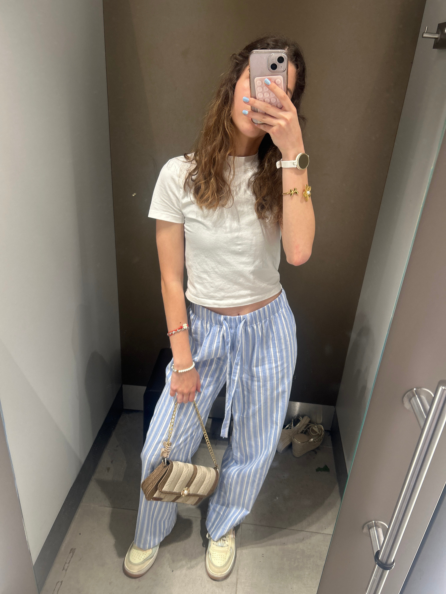A young woman with curly brown hair is taking a mirror selfie in a fitting room. She is wearing a white t-shirt and light blue and white striped pajama pants. She has a white smartwatch and several bracelets on her wrists, including a gold bracelet with a pearl and a red and white beaded bracelet. She is holding a small, striped handbag with a gold chain strap. She is wearing light-colored sneakers. In the background, there are grey and brown walls, a mirror, and a pair of gold wedge sandals on the floor. The fitting room appears to be in a retail store. The image does not provide any specific geographical context within the city of Ruse, Bulgaria.