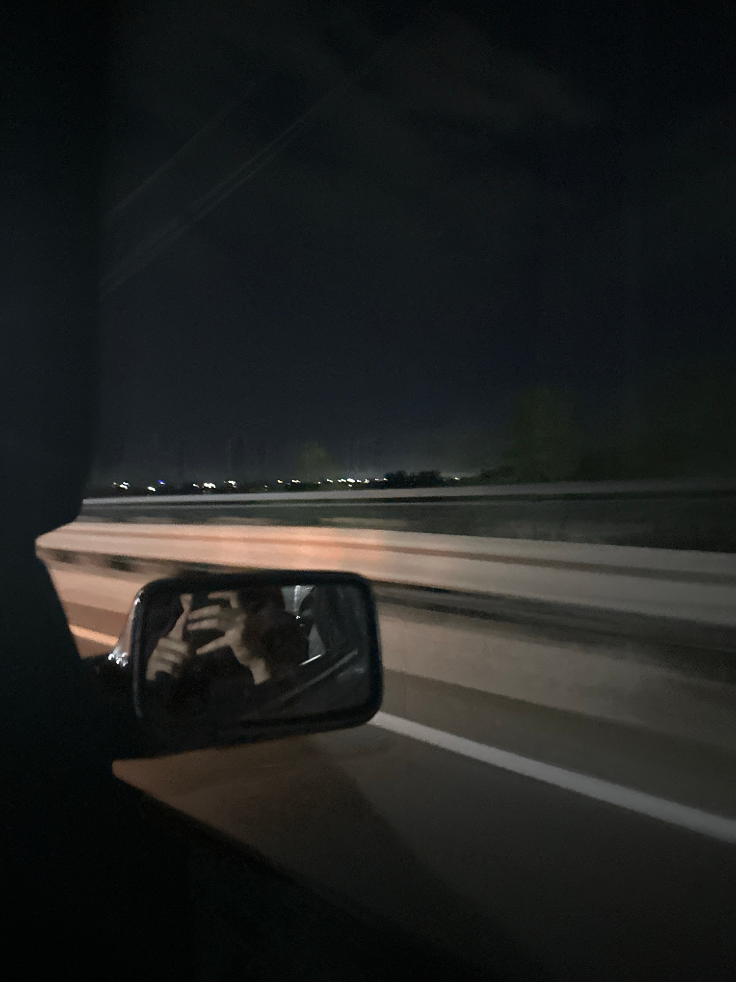 A low-angle, medium shot captures a nighttime view from inside a vehicle, looking out towards a highway. In the foreground, the dark silhouette of a car interior is visible on the left. A side-view mirror reflects a person holding a phone, appearing to take a selfie. The mirror is positioned on the left side of the frame. The highway stretches into the distance, illuminated by distant lights and a white line marking the lane. The sky is dark and clear. The scene is consistent with a view from a vehicle traveling on a highway near Kostanay, Kazakhstan, given the presence of highway infrastructure and the typical nocturnal lighting patterns seen in urban and suburban areas.