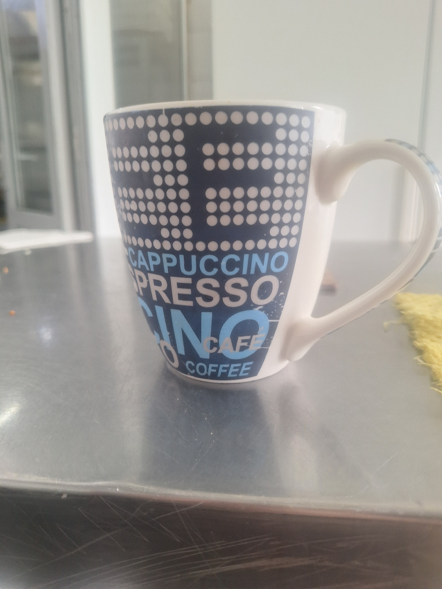 A white ceramic mug with a blue and white dot pattern and text is displayed on a brushed metal surface. The text on the mug reads "CAPPUCCINO", "ESPRESSO", "INO", "CAFE", and "COFFEE". The mug is positioned at a slight angle, with its handle visible on the right side. In the blurred background, a bright, possibly reflective surface, and a white structure with vertical elements are present. The metal surface shows some scratches and smudges, indicating it might be a table or countertop. The overall environment appears to be indoors, possibly in a kitchen or cafe setting, but no specific location in Shymkent, Kazakhstan, can be identified from the image alone.