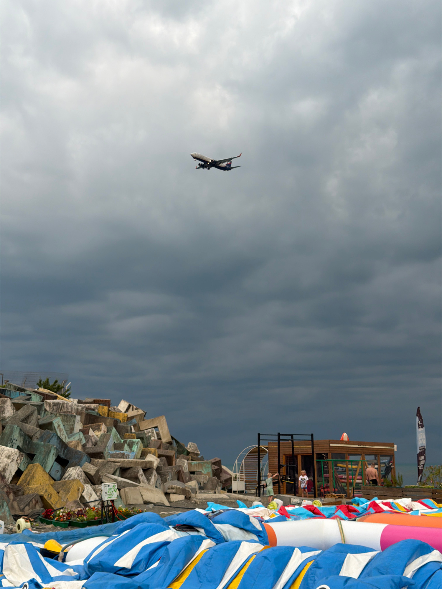 The image depicts a surreal, cloudy sky with a dramatic, dark undertone. In the distance, a small airplane is captured mid-flight, its dark silhouette contrasting with the lighter clouds. To the right, a vibrant, colorful collection of kites and kiteboarding gear is piled high, with a hint of a building structure behind them, possibly a kite school or shop. A few people are visible near the building, engaged in various activities, though their details are indistinct due to the distance and resolution.
The composition is unusual, with the right side of the image oriented vertically, showing a chaotic but intriguing arrangement of concrete blocks, suggesting an artistic installation or perhaps a construction site. The colors of the kites, particularly the blues, whites, and oranges, create a stark and lively contrast with the muted sky and the earthy tones of the concrete structures.
The overall impression is one of anticipation or transition. The dark, moody sky could indicate an approaching storm, while the colorful kites and the presence of people suggest an active atmosphere, possibly related to water sports or an event. The time of day appears to be late afternoon or early evening, given the lighting and cloud cover. The location, identified as Sirius, Russia, adds a specific geographical context to this eclectic scene. There is no discernible text within the image.