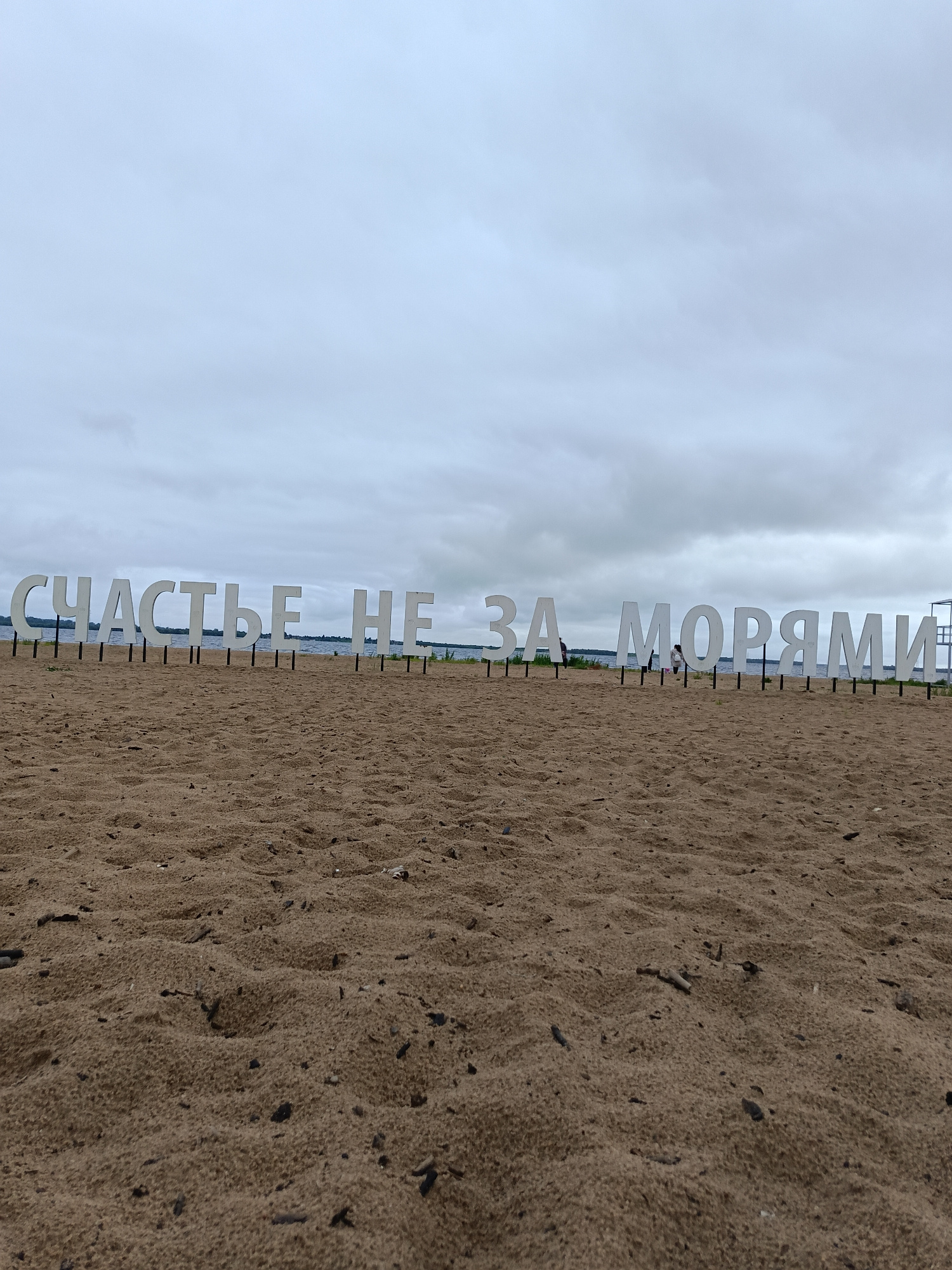 The image captures a beach scene with large, white, three-dimensional letters spelling out "СЧАСТЬЕ НЕ ЗА МОРЯМИ" (Happiness is not beyond the seas) in Russian. The letters are supported by black poles and are placed on a sandy beach. In the background, a body of water, likely the Severnaya Dvina River or White Sea, is visible under a cloudy sky. A few indistinct figures can be seen near the letters. The foreground is dominated by the textured sand of the beach. The location is the Arkhangelsk embankment in Arkhangelsk, Russia.