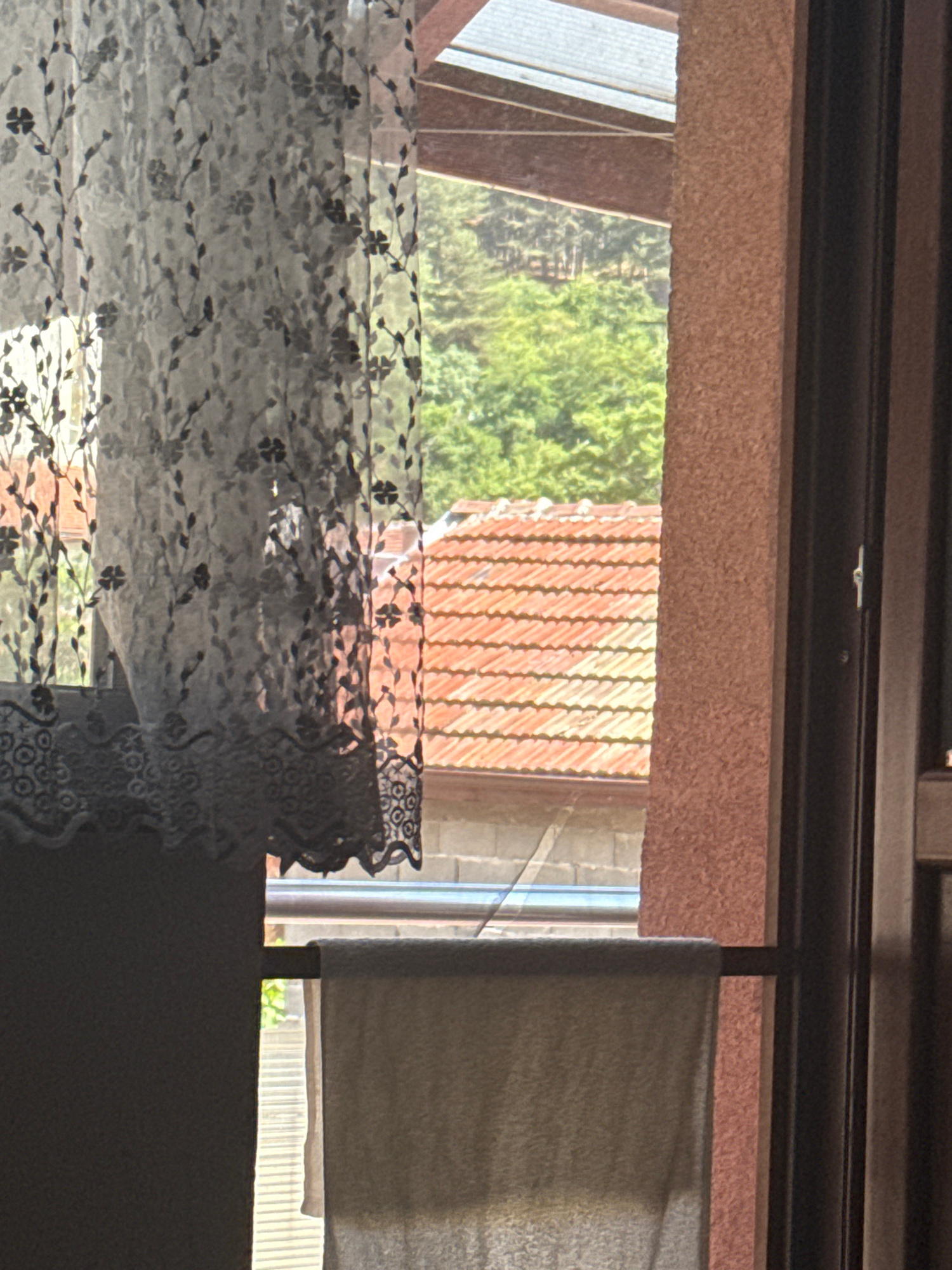 A close-up, eye-level shot captures a view through a window. In the foreground, a sheer, white lace curtain with a floral pattern drapes across the left side of the frame. Below the curtain, a thick, darker fabric hangs, obscuring the lower portion of the window. The middle ground reveals a residential scene outside: a section of an orange-tiled roof is visible, with a hint of a wall structure beneath it. Beyond that, lush green trees form a dense backdrop, suggesting a somewhat rural or suburban setting. The far right of the image shows a textured, salmon-colored wall and a dark vertical frame, likely part of the window or door structure. A light-colored towel is hung on a horizontal rod across the lower part of the window. The lighting suggests it is daytime, with sunlight illuminating the scene from the right. The image does not contain any people or indicate specific actions. The view is consistent with a residential setting in Dupnitsa, Bulgaria, possibly overlooking a garden or neighboring houses with abundant vegetation.